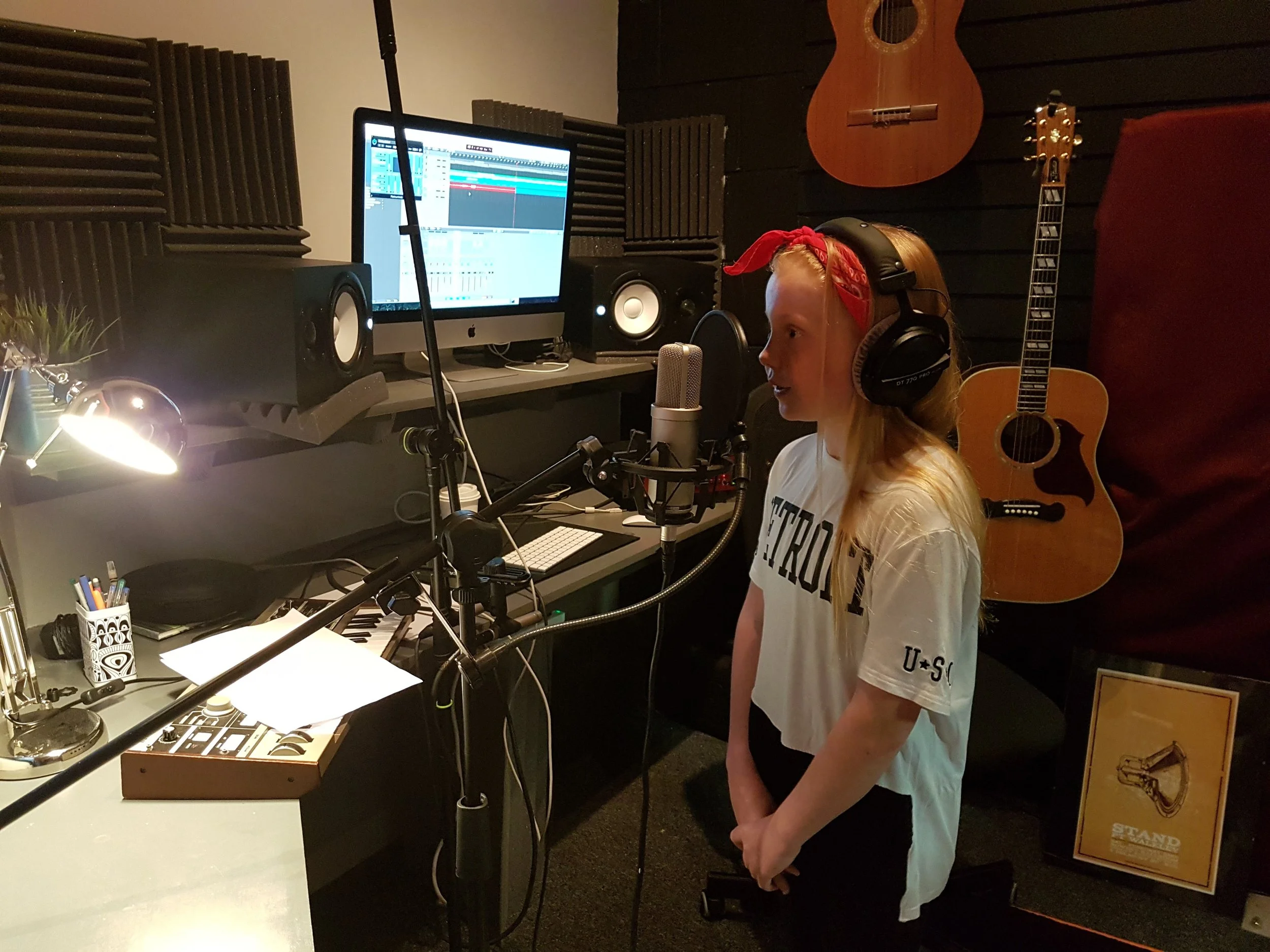 Person wearing headphones and standing at a microphone in a recording studio with acoustic foam, a computer with sound mixing software, and guitars on the wall.