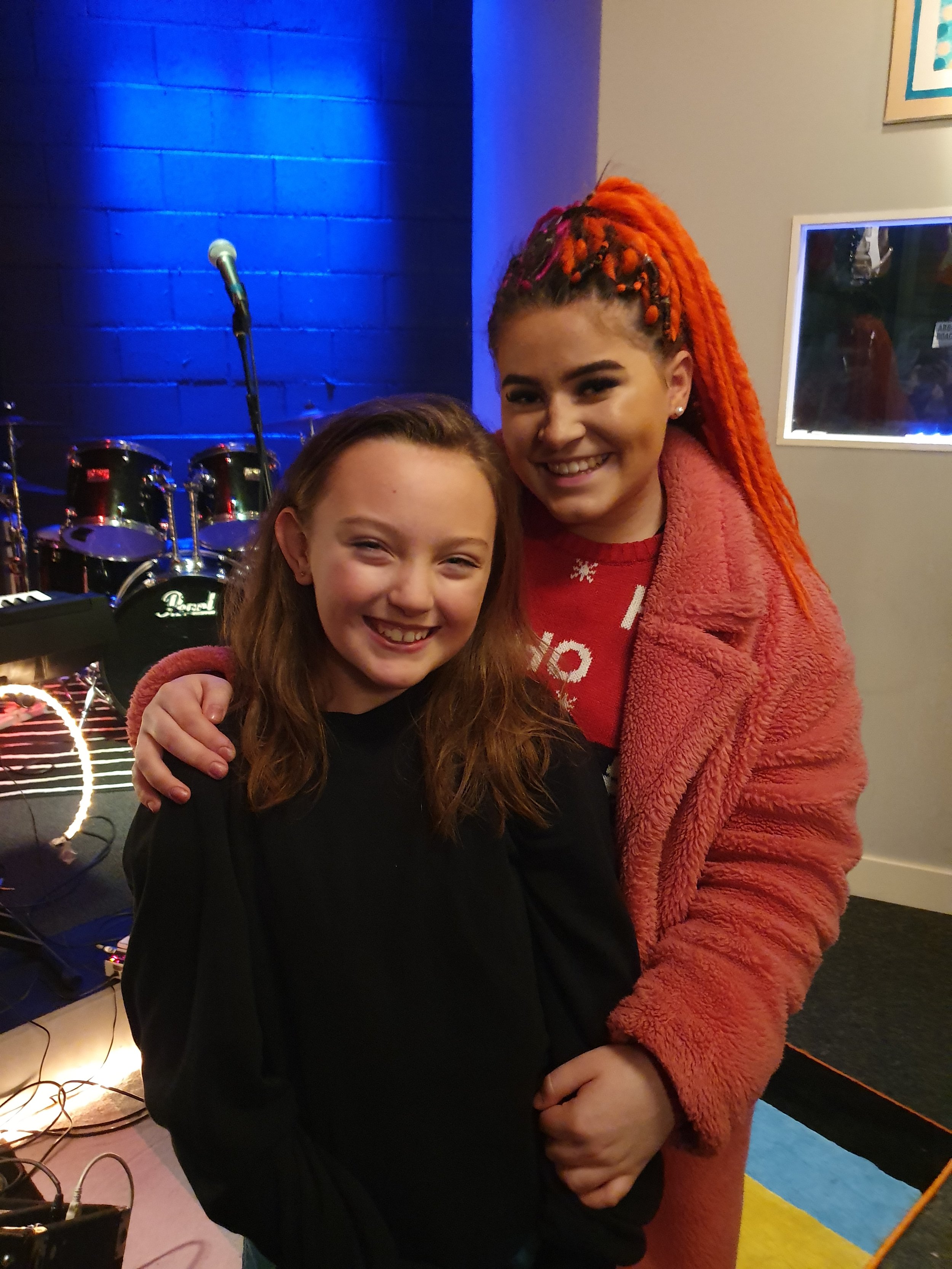 Two people smiling in a music studio with microphones and drums in the background, one wearing a pink coat.