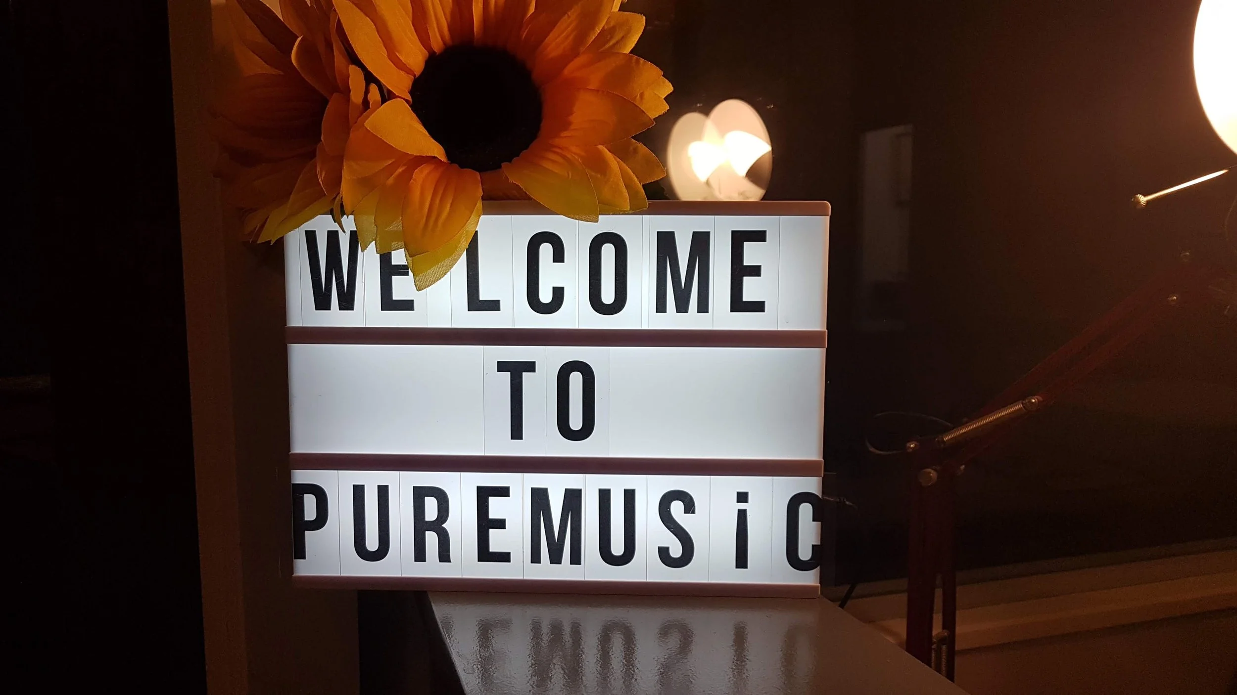 Illuminated sign with 'WELCOME TO PURE MUSIC,' sunflower decoration, and lamps in background.