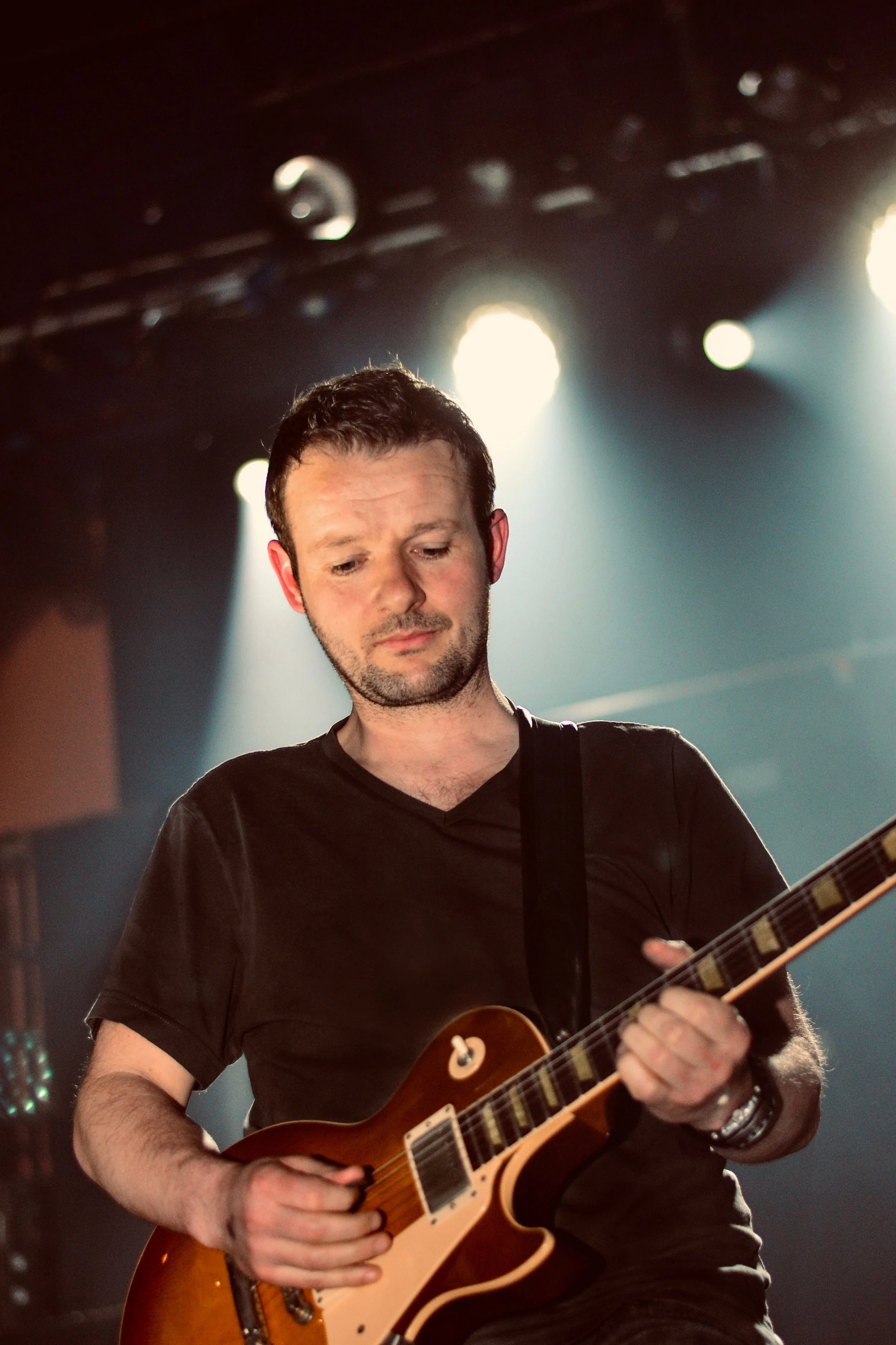 Musician playing an electric guitar on stage with spotlights in the background.