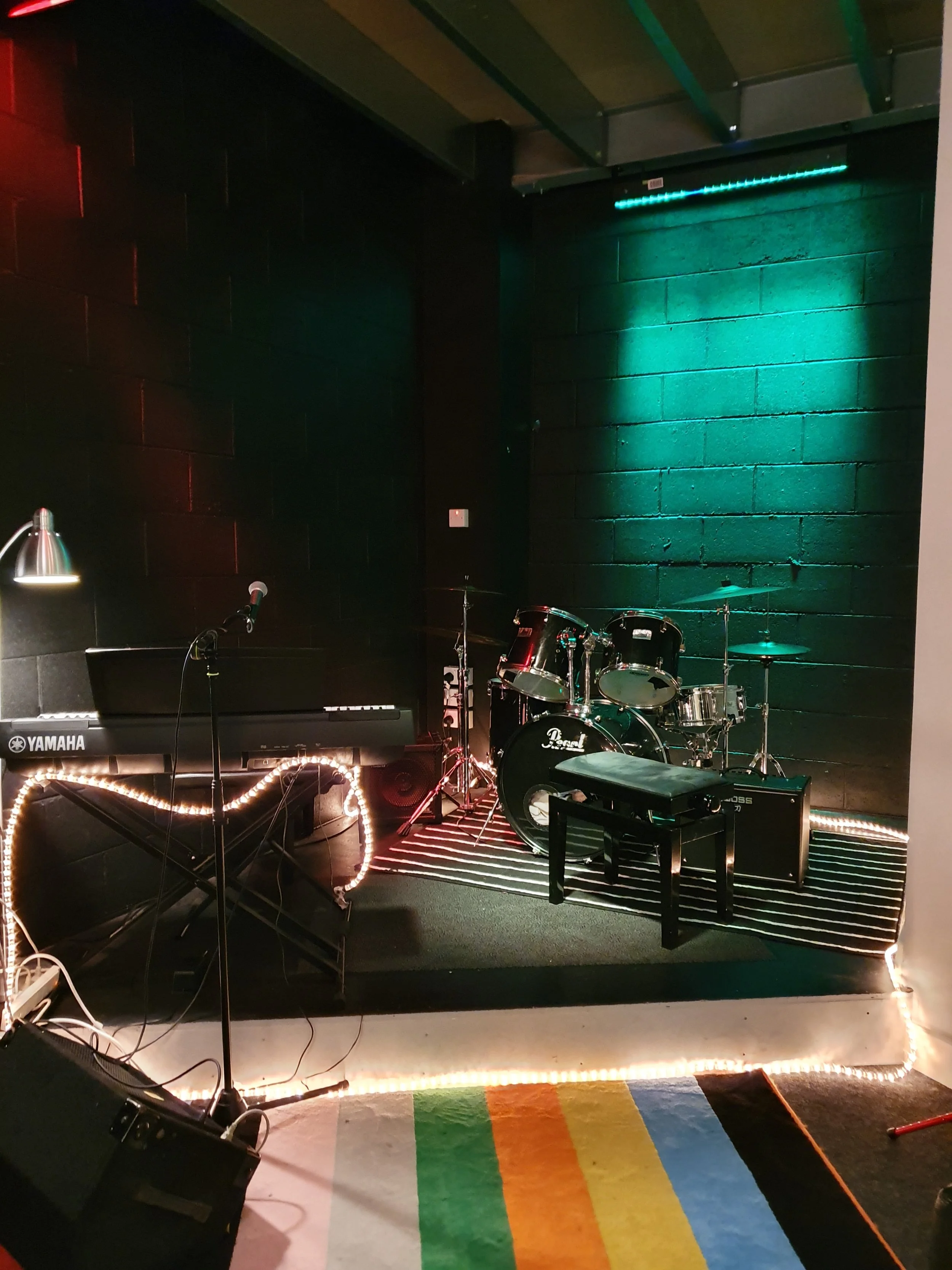 Small music stage with a drum set, keyboard, microphone, and amplifier, illuminated by colorful lights and surrounded by striped patterns on the floor.