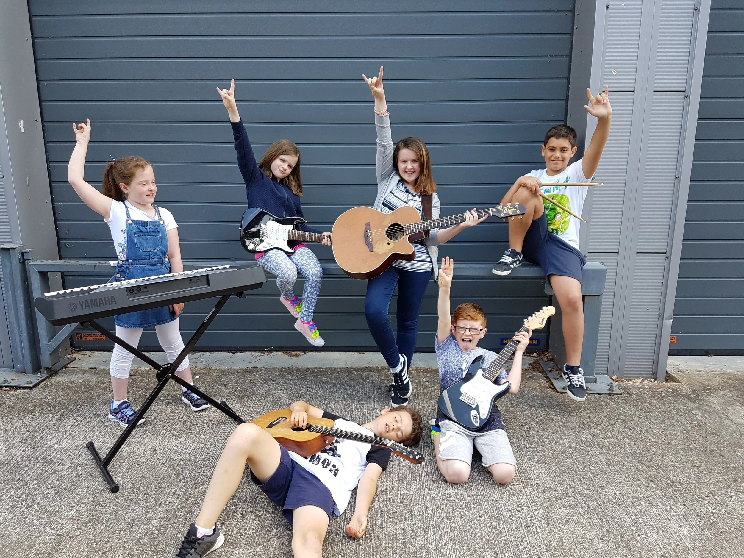 Group of children posing with musical instruments in front of a gray garage. One plays a keyboard, another holds an electric guitar, one has an acoustic guitar, a child sits with drumsticks, and another lies on the ground with a guitar. All raise hands in a rock gesture.