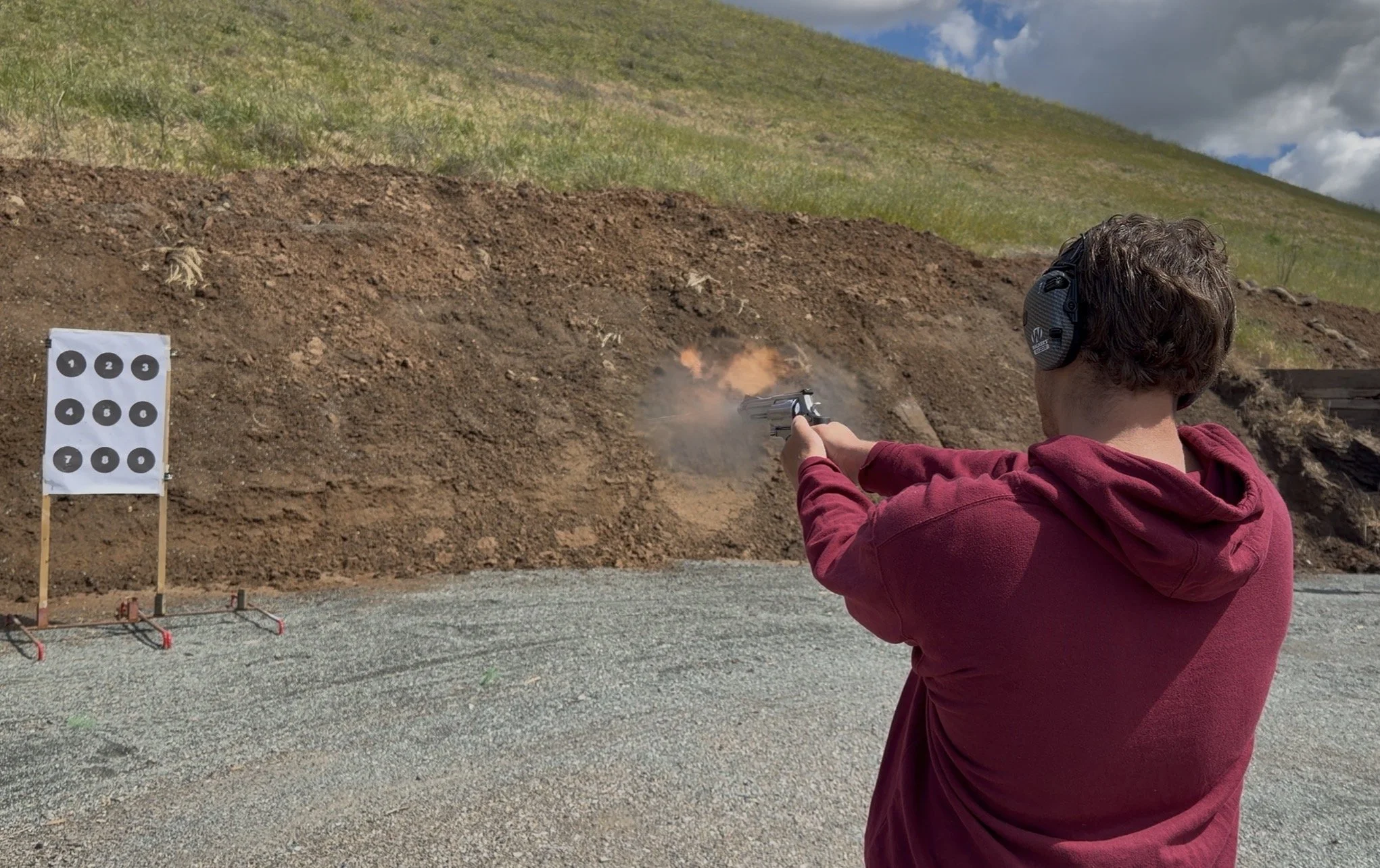 A person wearing a red hoodie and protective ear muffs is firing a handgun at a shooting range. There is a target with numbered circles on a stand to the left.