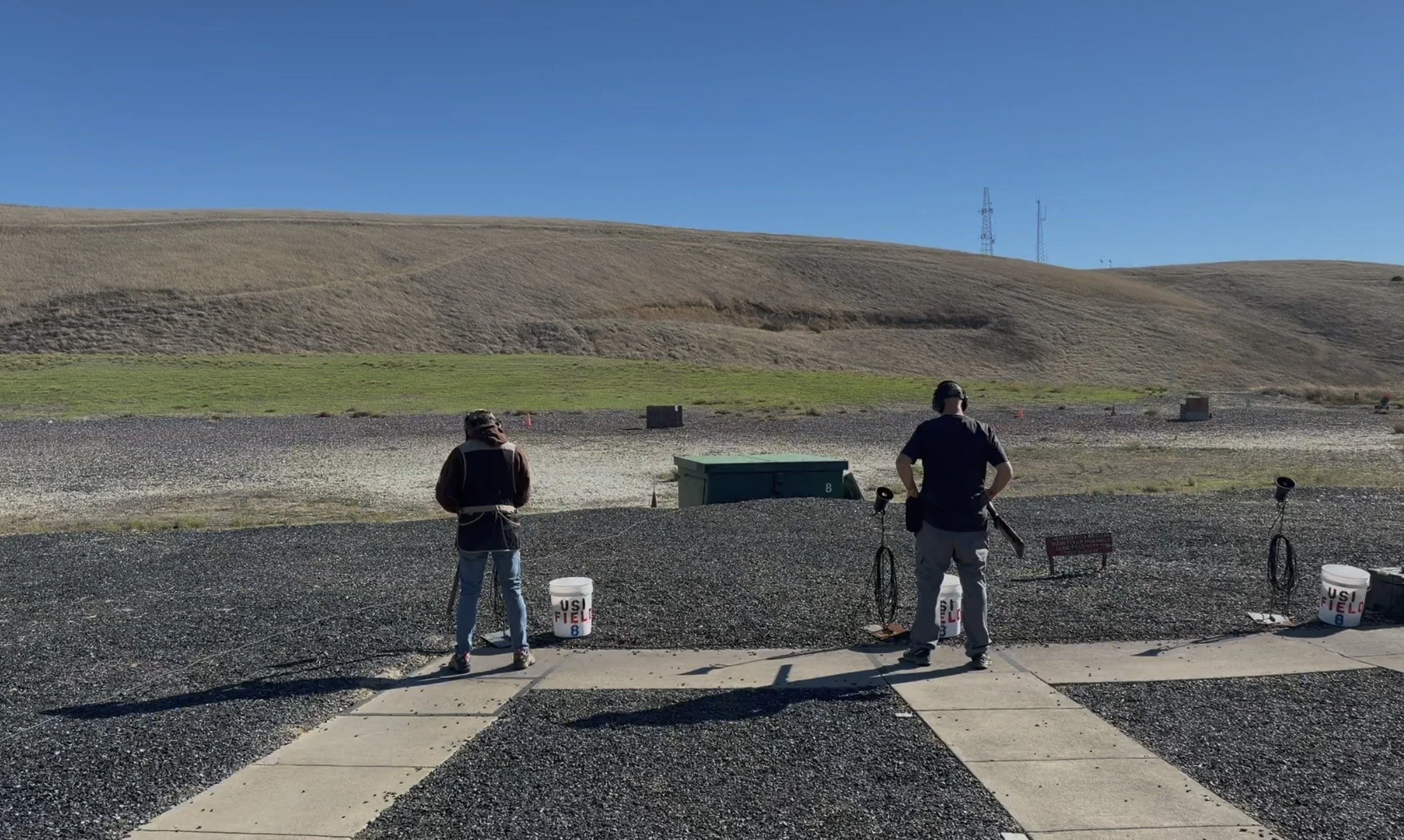 Two people are shooting at an outdoor firing range with hills and clear blue sky in the background. They are standing behind safety barriers, with targets and equipment in front of them. Buckets labeled 'USF FIELD 8' are on the ground.