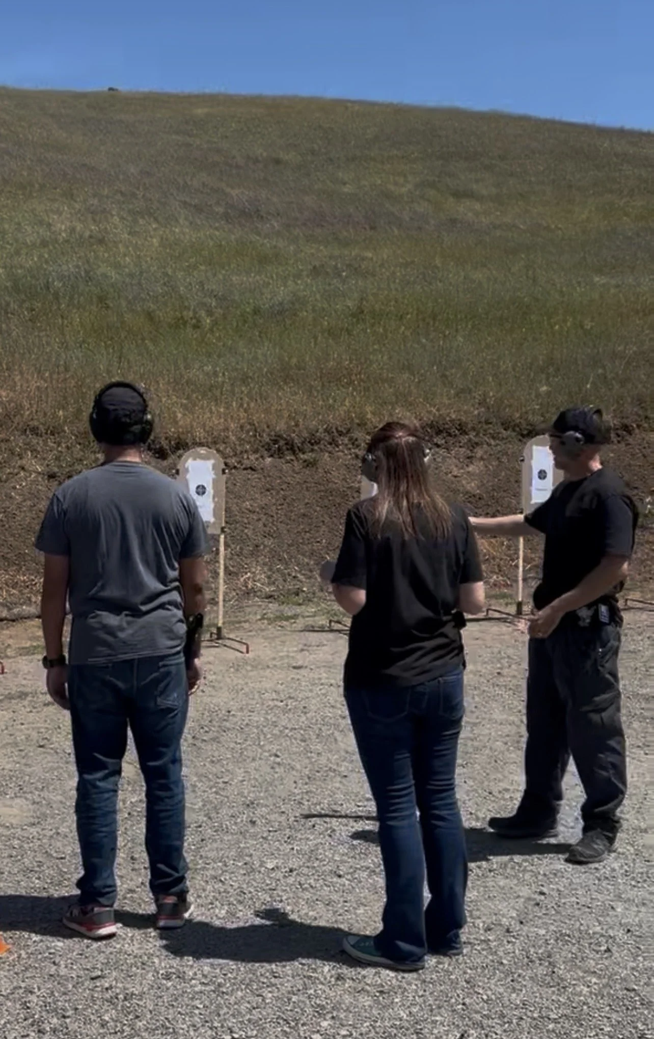 Three people at an outdoor shooting range aiming at targets mounted on posts. Two men and one woman, all wearing ear protection, are standing on a gravel surface with a grassy hillside in the background and a clear blue sky above.
