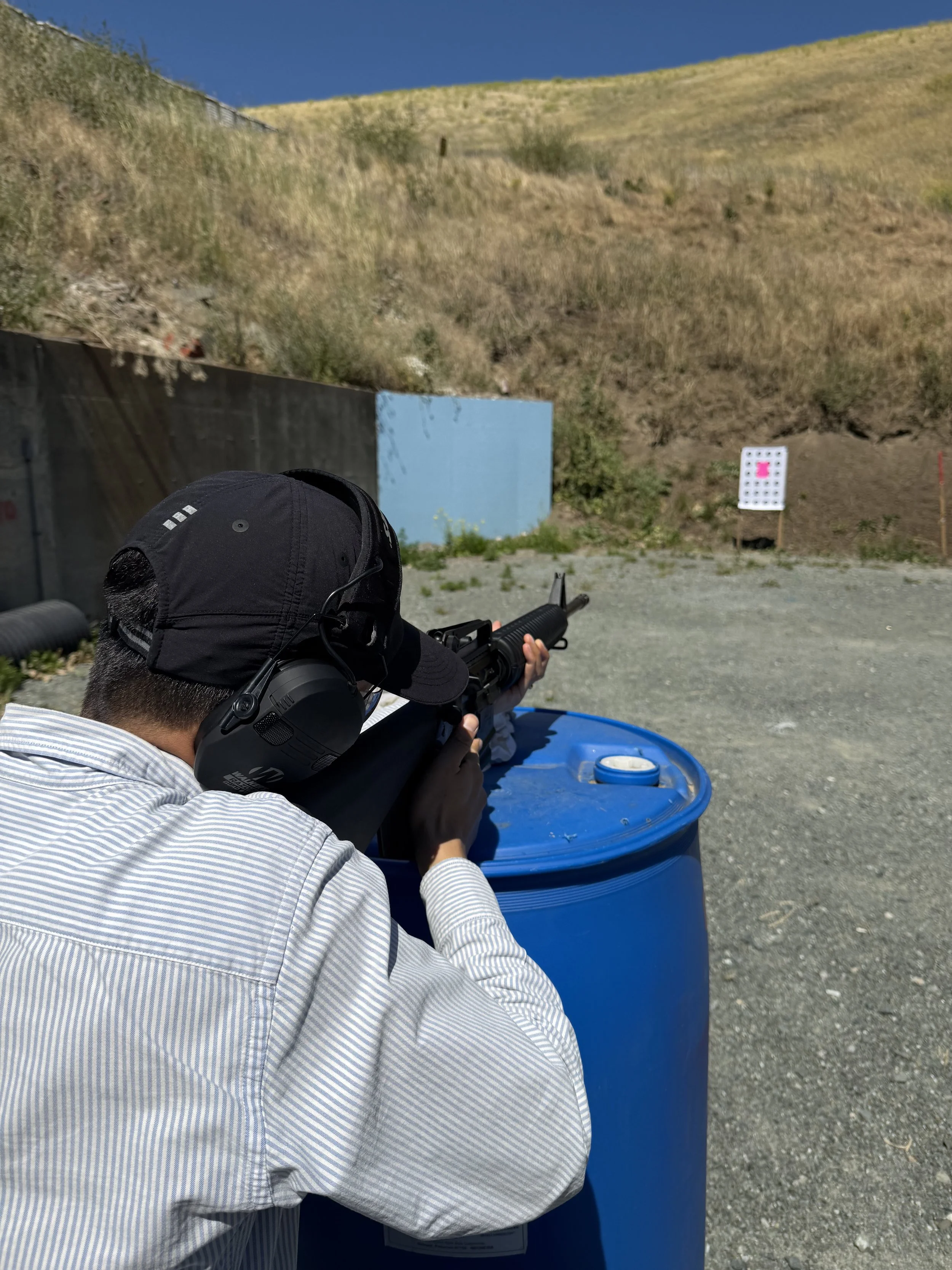 A person wearing a white and blue striped shirt, black headphones, a black hat, and glasses is aiming a rifle at a target during shooting practice outdoors on a sunny day. The target is visible in the distance against a hillside.
