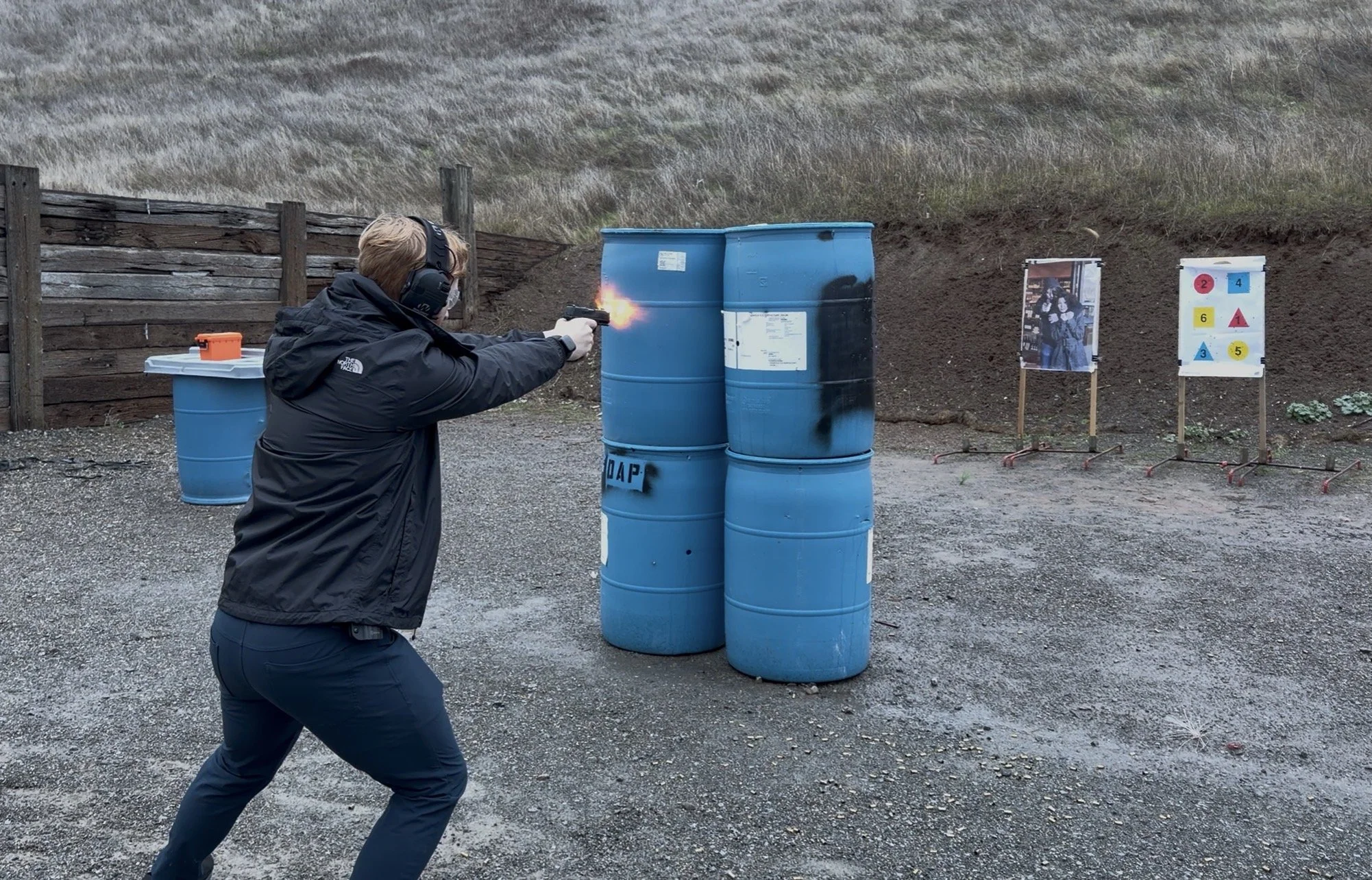 A person wearing a black jacket and headset is firing a gun in an outdoor shooting range. They are aiming at a paper target that has a photo of three people. There are several blue barrels nearby and a wooden fence in the background.