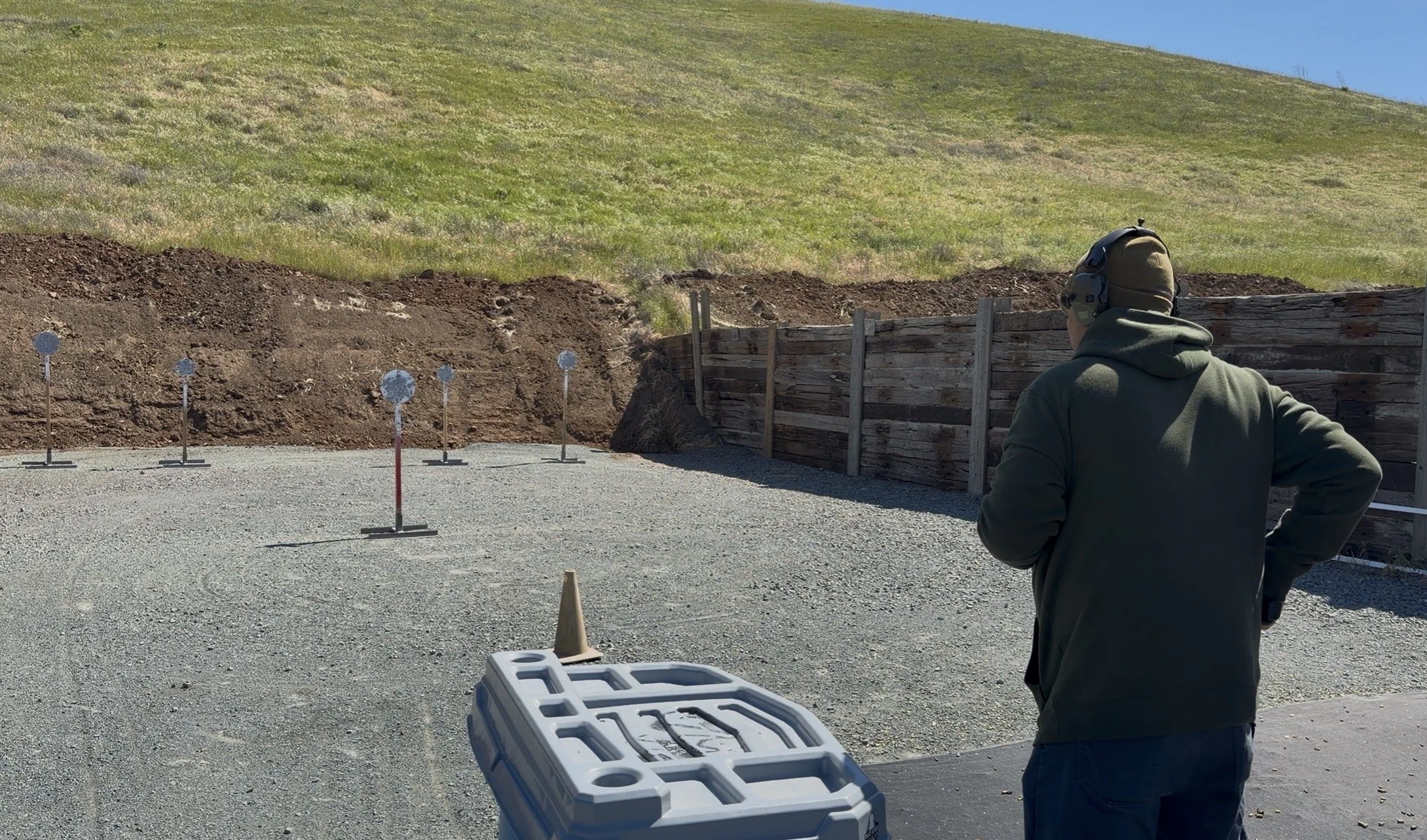A person at a shooting range standing outdoors, wearing a hoodie and headphones, facing targets set up against a wooden barrier with a hillside in the background.