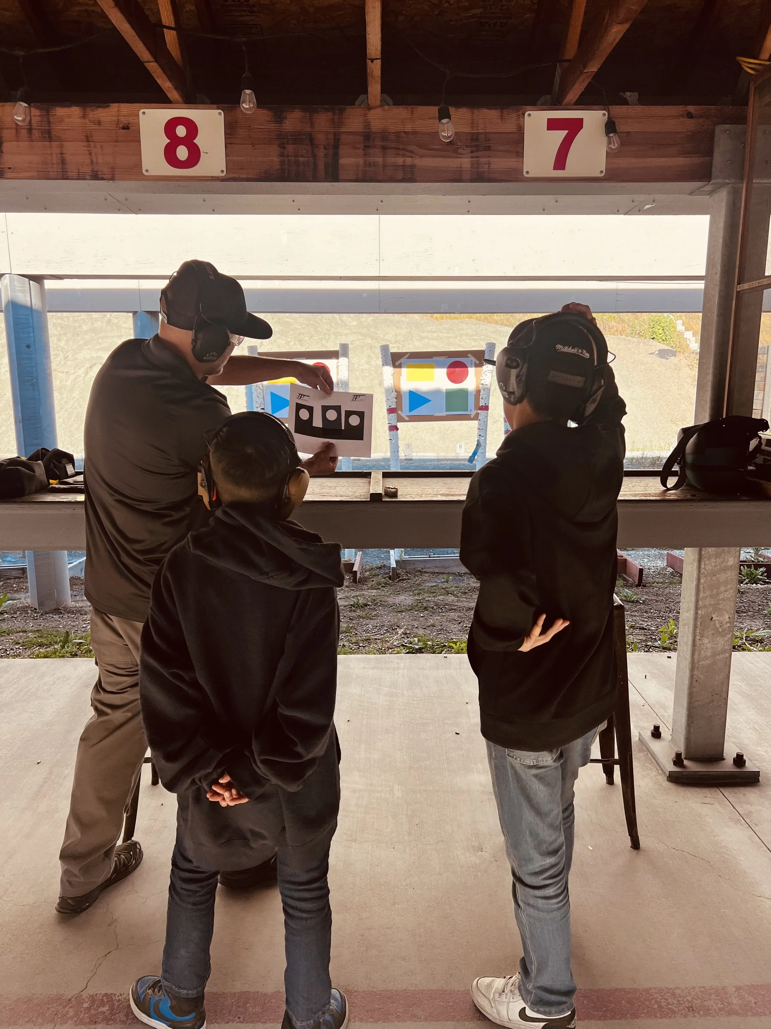 Four people standing at an outdoor shooting range, two adults and a child, observing targets on the other side. The range has signs labeled '8' and '7' above, and target boards with colored shapes. The people are wearing ear protection and casual clothing.