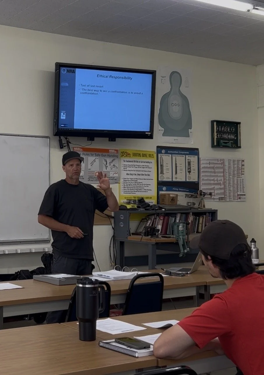A classroom or training room with a man standing at the front, giving a lecture or presentation. On the wall behind him is a large screen displaying a slide titled 'Ethical Responsibility.' There are two students seated at tables listening, with one of them taking notes and the other attentively listening.