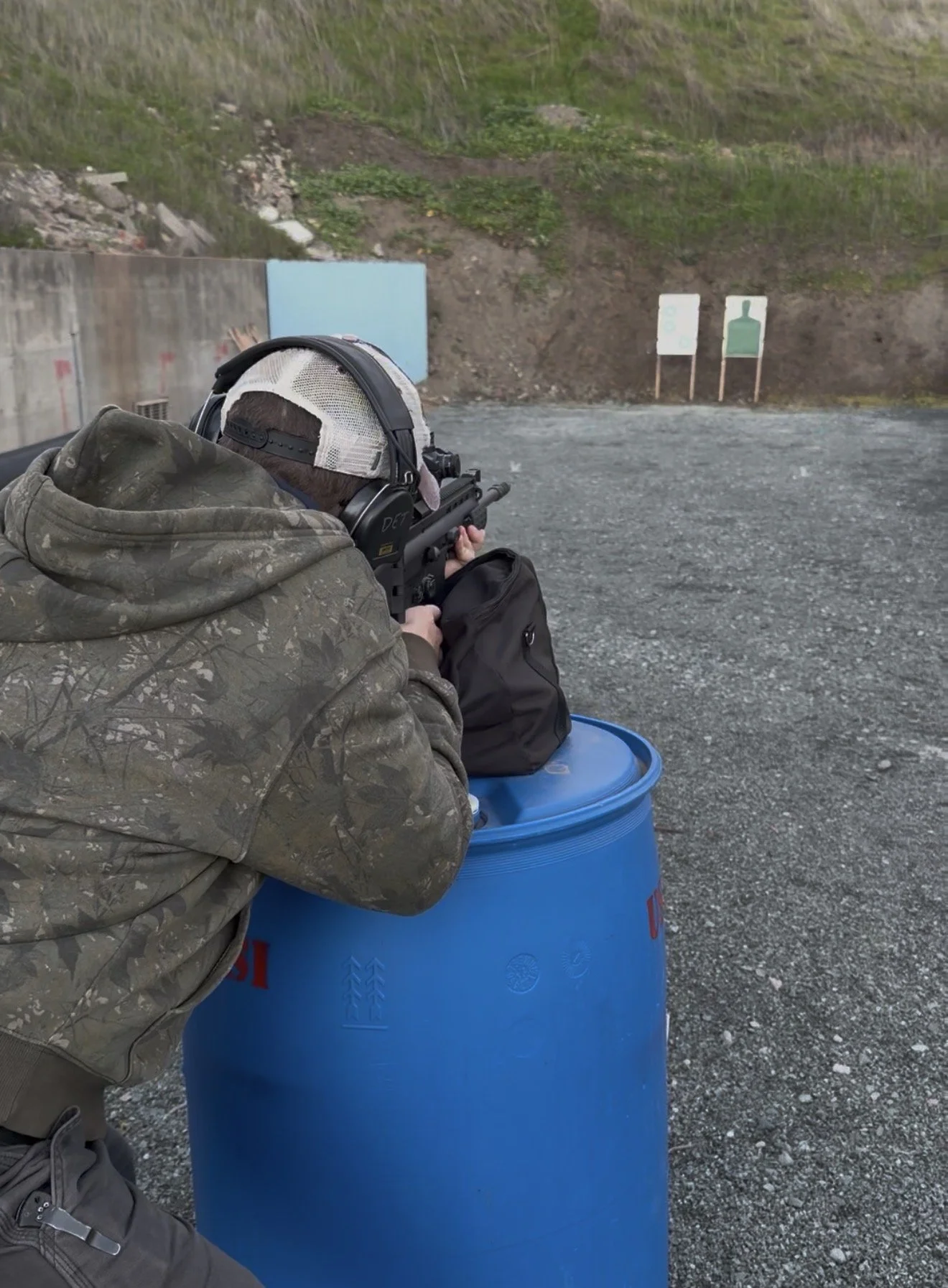 Person in camouflage jacket and baseball cap aiming a firearm at shooting targets outdoor, with gravel ground and green hillside background.