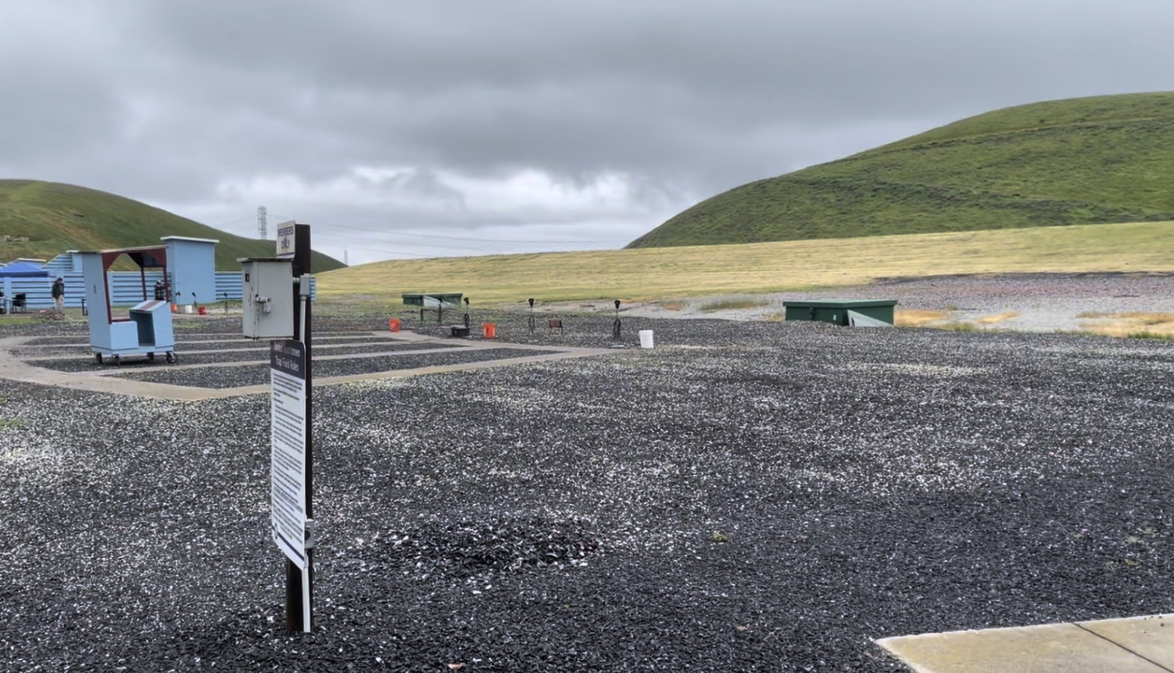 Gravel parking lot near green hills with cloudy sky. Several outdoor electrical and utility boxes, a green bench, and a sign in the foreground. Trap and Skeet field.