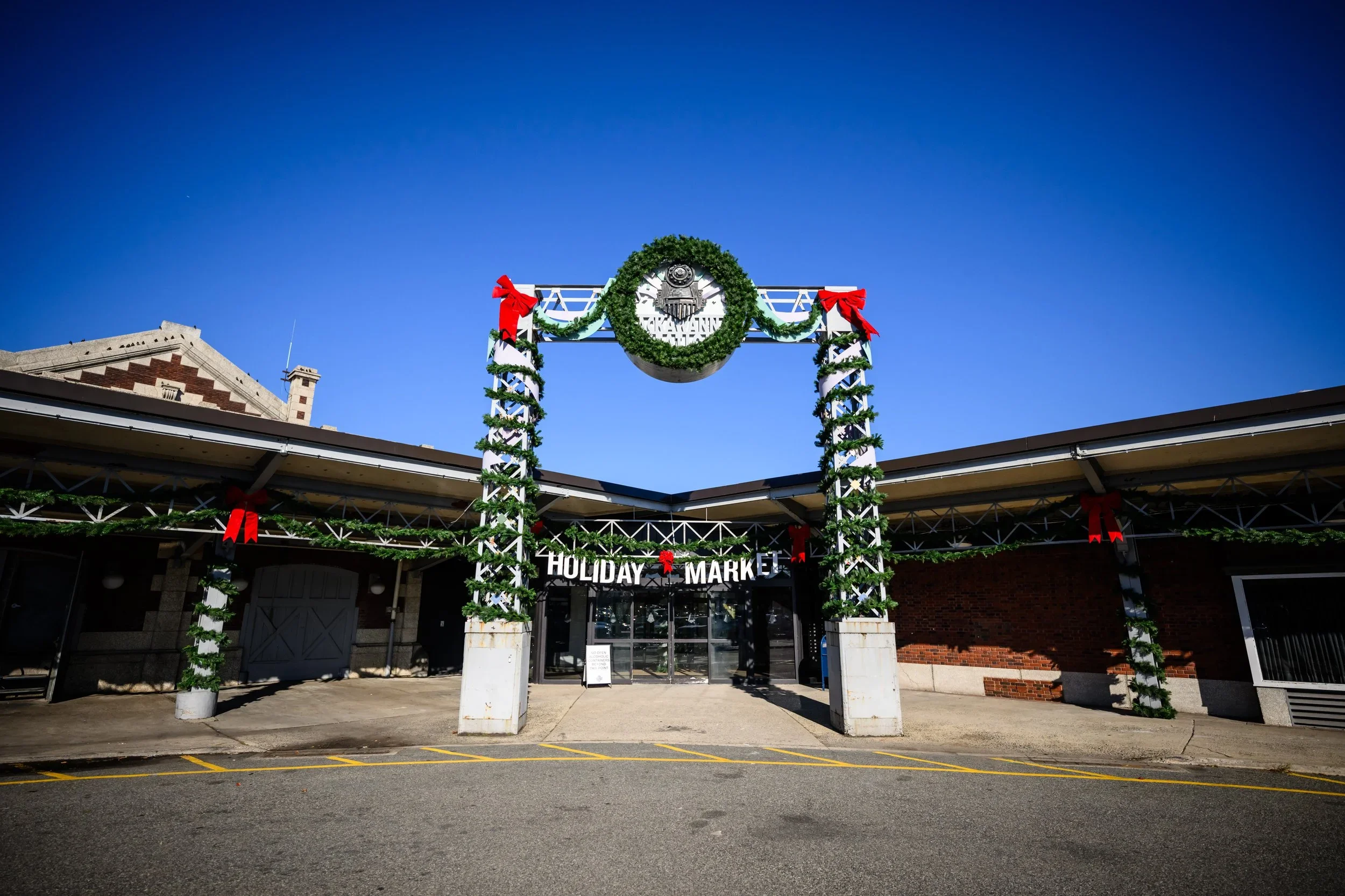 Decorated entrance to a holiday market with garlands, red bows, and a wreath with a train logo, under a clear blue sky.