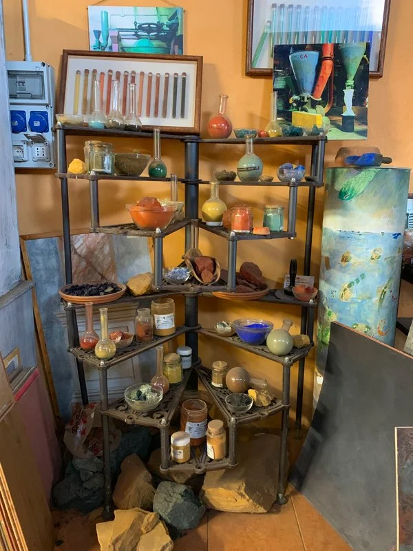 A display of various colorful glass and ceramic laboratory glassware and minerals arranged on a black metal shelf, with paintings, rocks, and art supplies in the background.