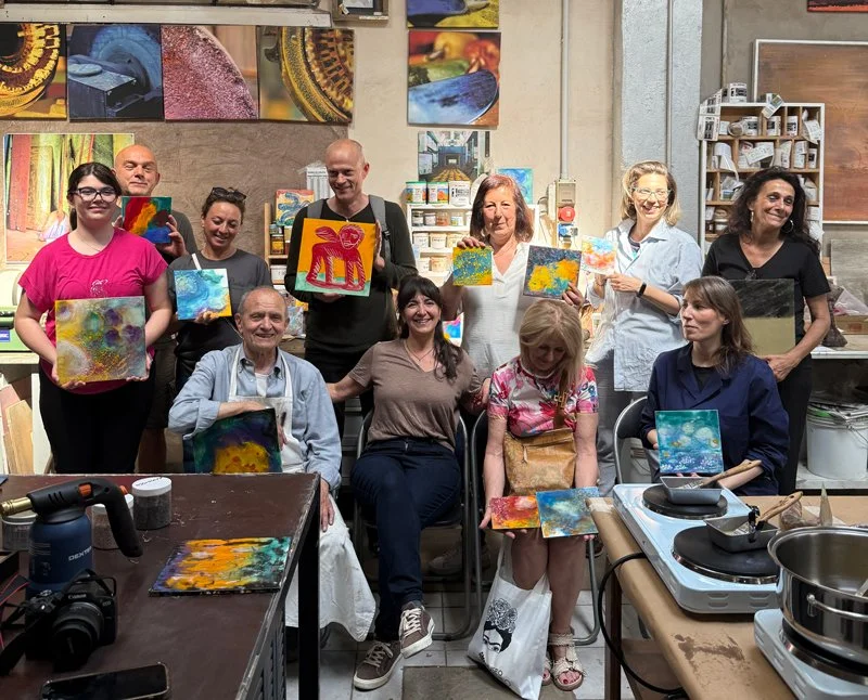 Group of people in an art workshop holding colorful abstract and animal-themed paintings, with art supplies and paintings on the wall in the background.