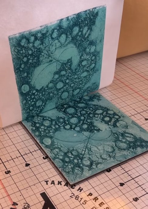 Close-up of a handmade art piece showing a patterned paper being peeled from a white surface, with a grid cutting mat underneath.