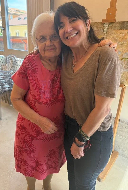Two women hugging and smiling inside a room with large windows, one older woman wearing a red dress with palm leaf pattern and glasses, the other younger woman wearing a brown t-shirt and dark jeans.