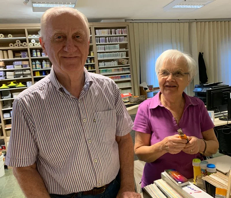 An elderly man and woman standing indoors in a room with shelves filled with various items, including paper rolls and storage boxes, and a table with books, pills, and a hot sauce bottle.