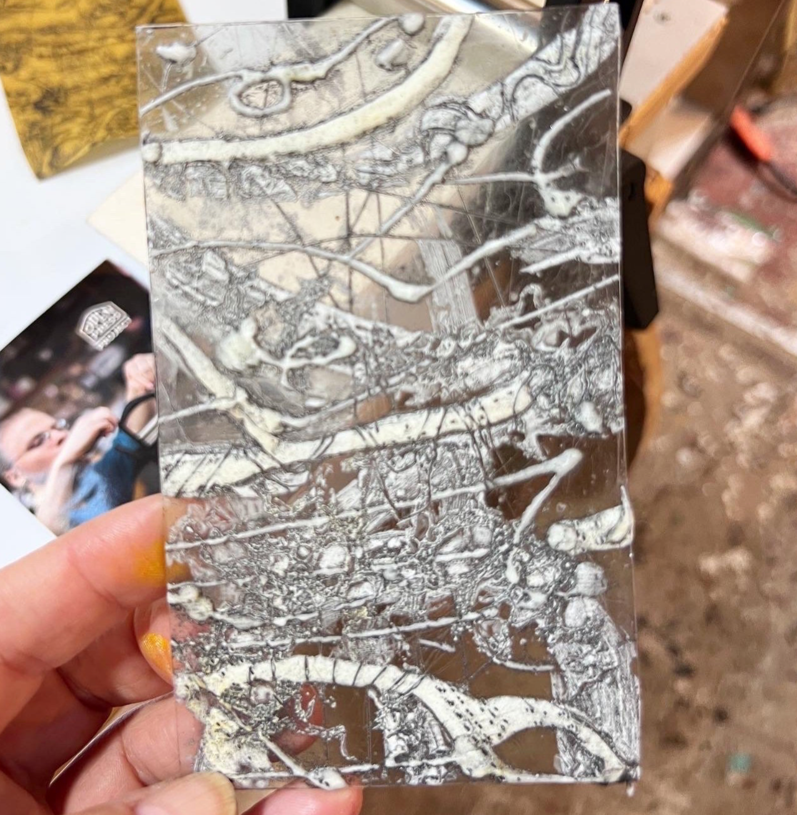 Hand holding a small rectangular sheet of glass with white paint splatters and streaks.