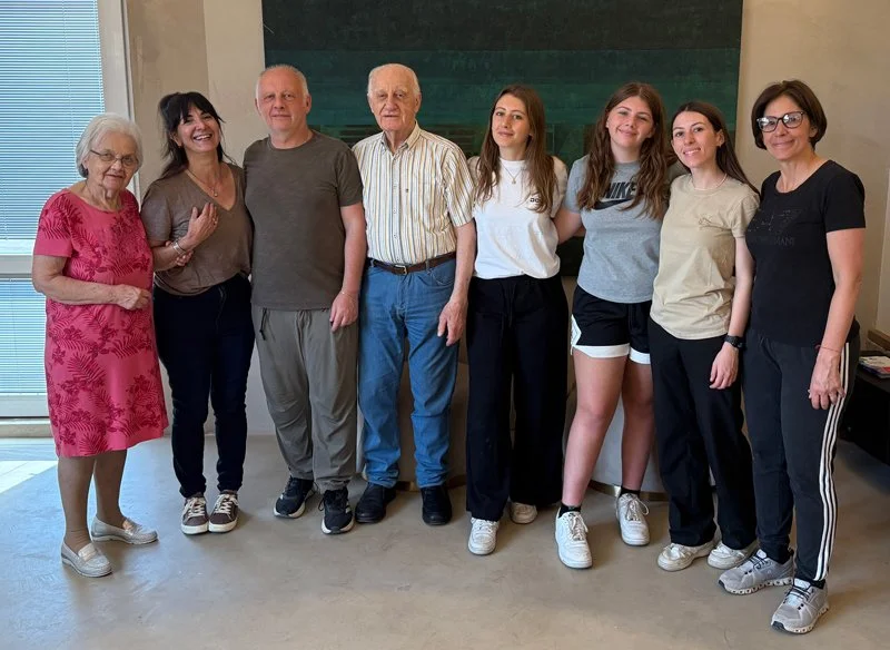 A group of eight people standing together indoors, smiling, with varying ages and casual attire.
