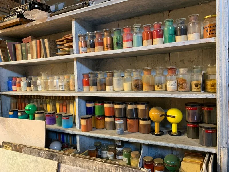A wooden shelf filled with glass jars containing various powders, pigments, and substances used for artwork, along with a few color calibration devices.