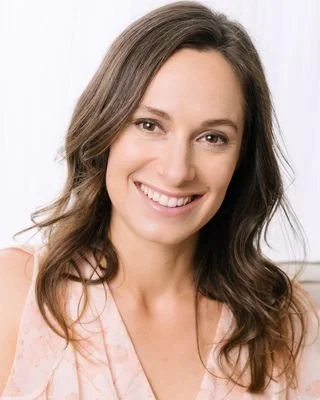 Smiling woman with brown hair, wearing a light-colored top, against a white background.