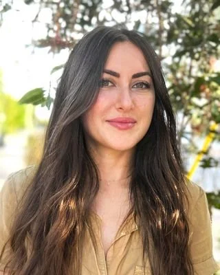 Woman with long brown hair smiling outdoors with blurred greenery in the background.