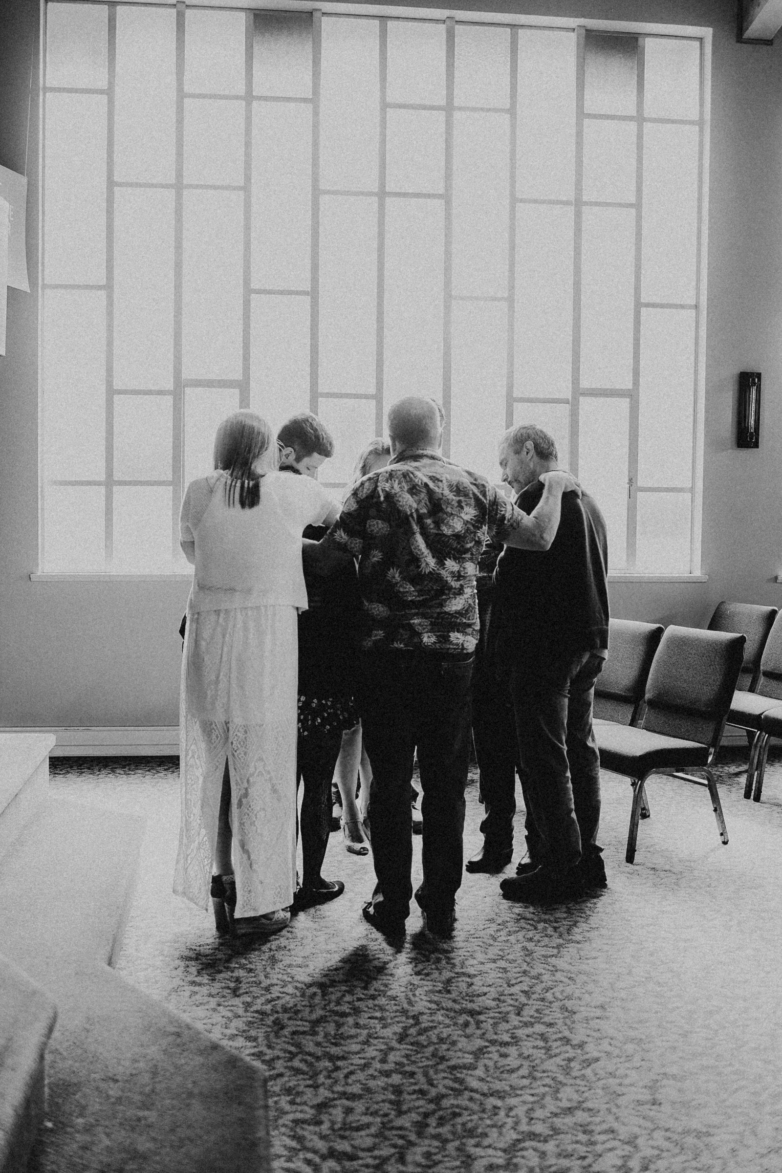 A group of six people stand closely together in a circle inside a well-lit room with large windows in the background. The individuals are engaged in conversation and appear to be sharing an intimate or meaningful moment. Some are wearing masks, and the group consists of both men and women of varying ages.