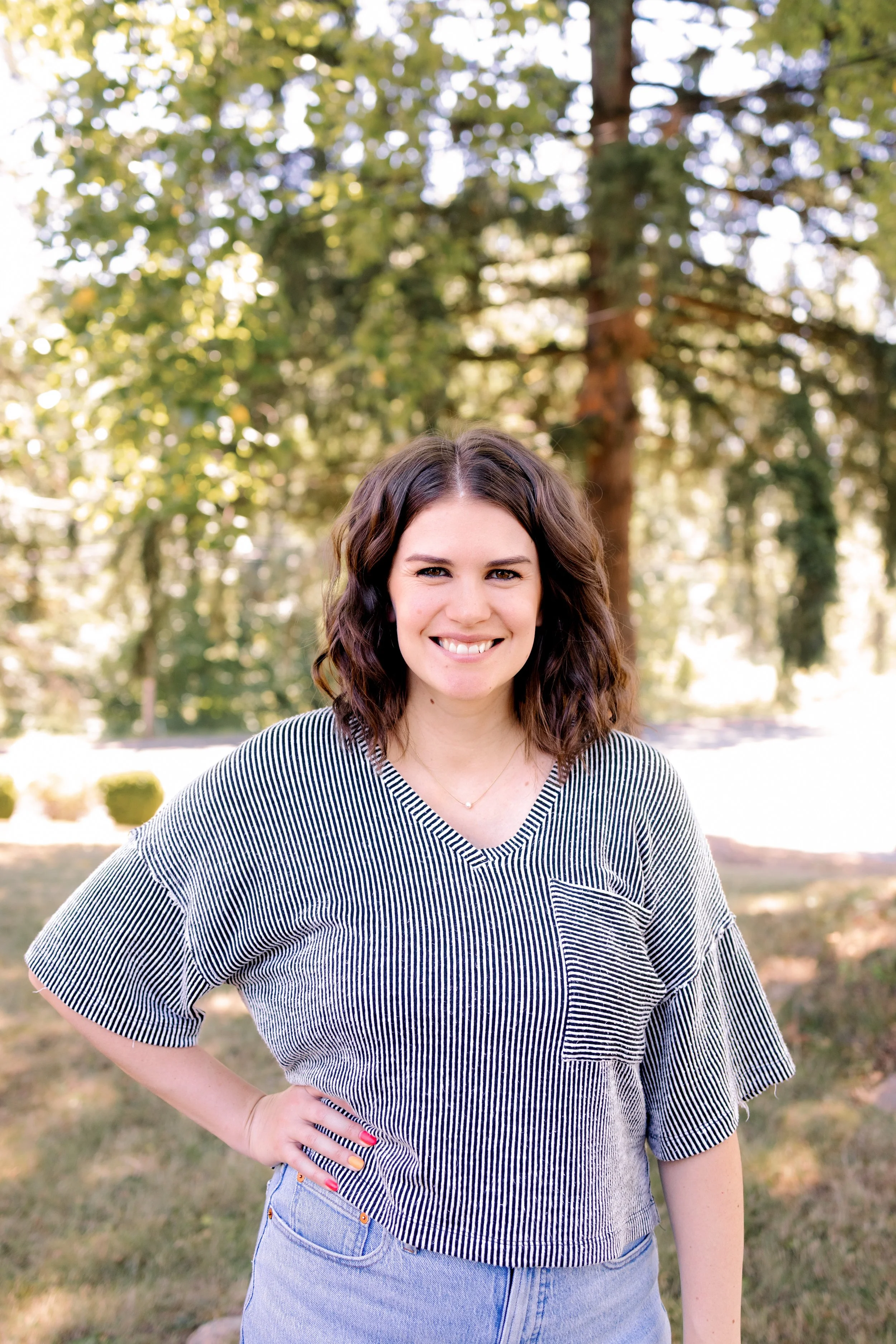 A young woman standing outdoors in a park with trees in the background, smiling and wearing a striped short-sleeve shirt and light blue jeans.