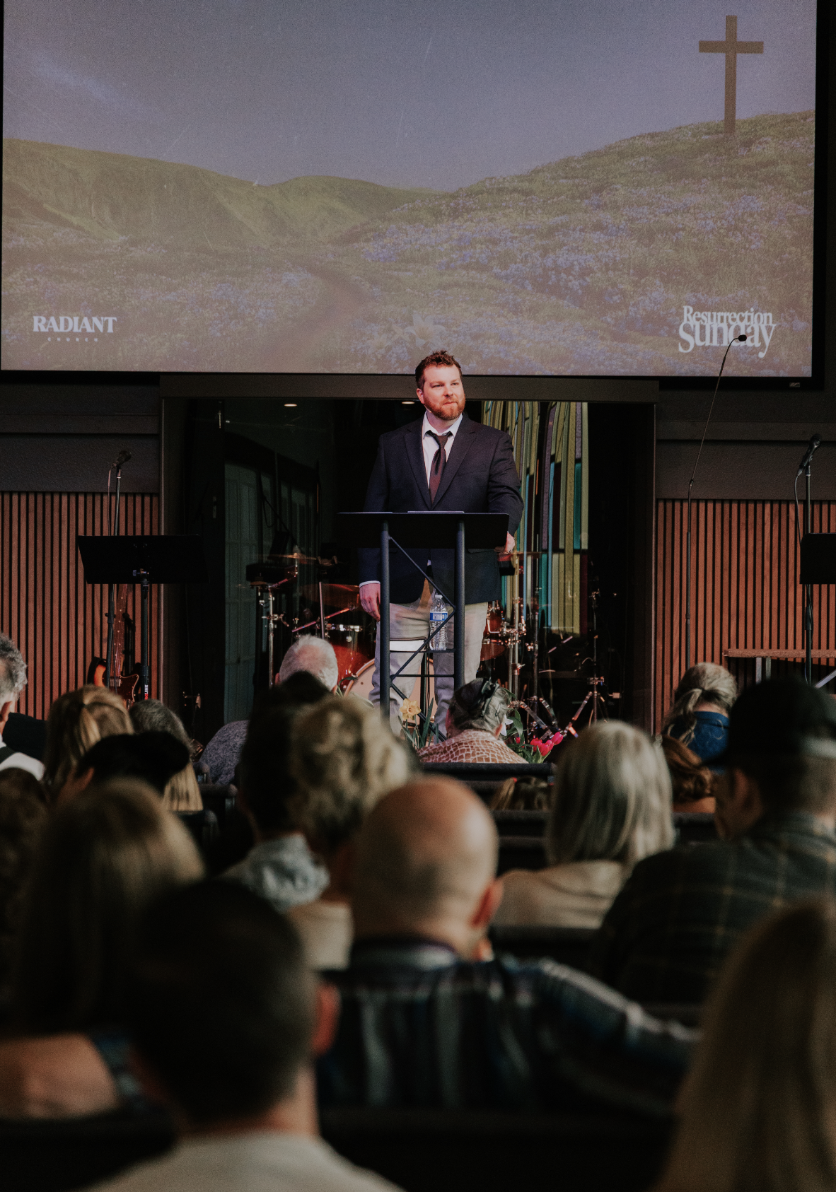 Man speaking at a podium during a church service or event, with a cross on a hill and a large screen in the background displaying a landscape with purple flowers and the words 'Resurrection Sunday'. Congregation seated in front.