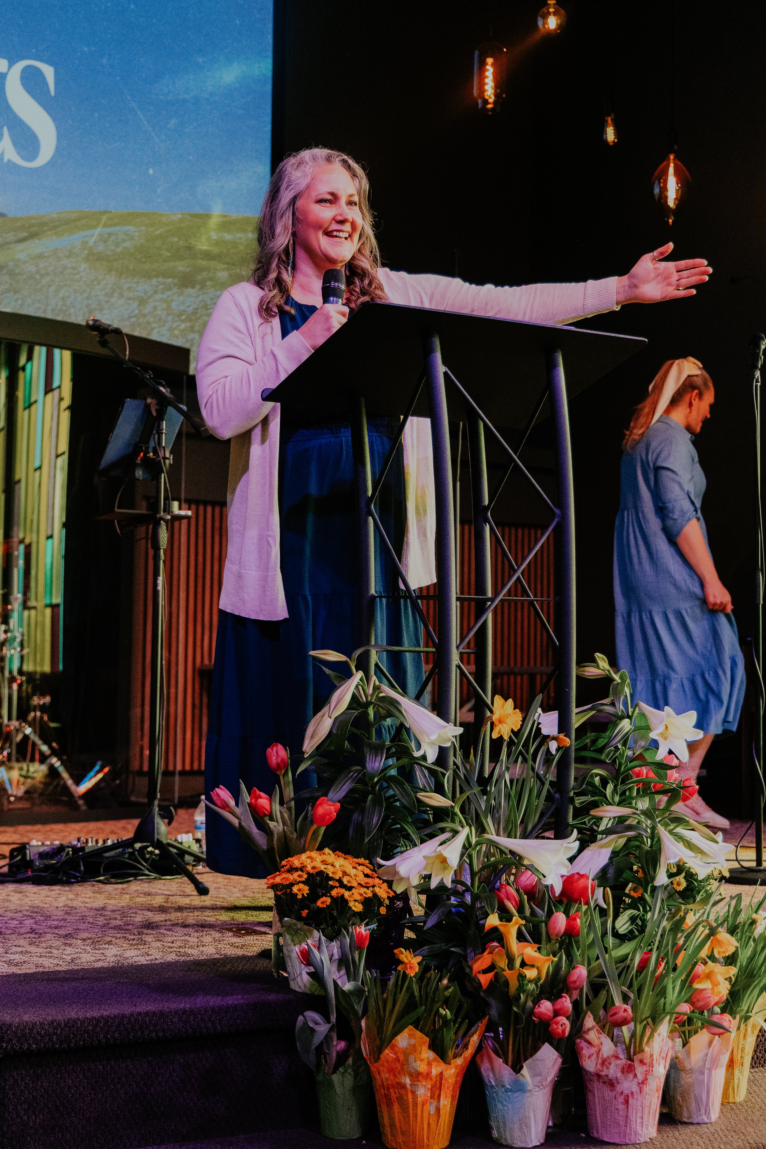A woman speaking at a stage with a microphone in hand, gesturing outward. She is dressed in a pink cardigan over a dark dress and has long, wavy gray hair. The stage is decorated with numerous colorful flower arrangements at the front, including lilies, tulips, and chrysanthemums, wrapped in decorative paper. In the background, another woman in a blue dress with a white headscarf is standing, facing downwards. The scene is set indoors with dim lighting and hanging light fixtures.