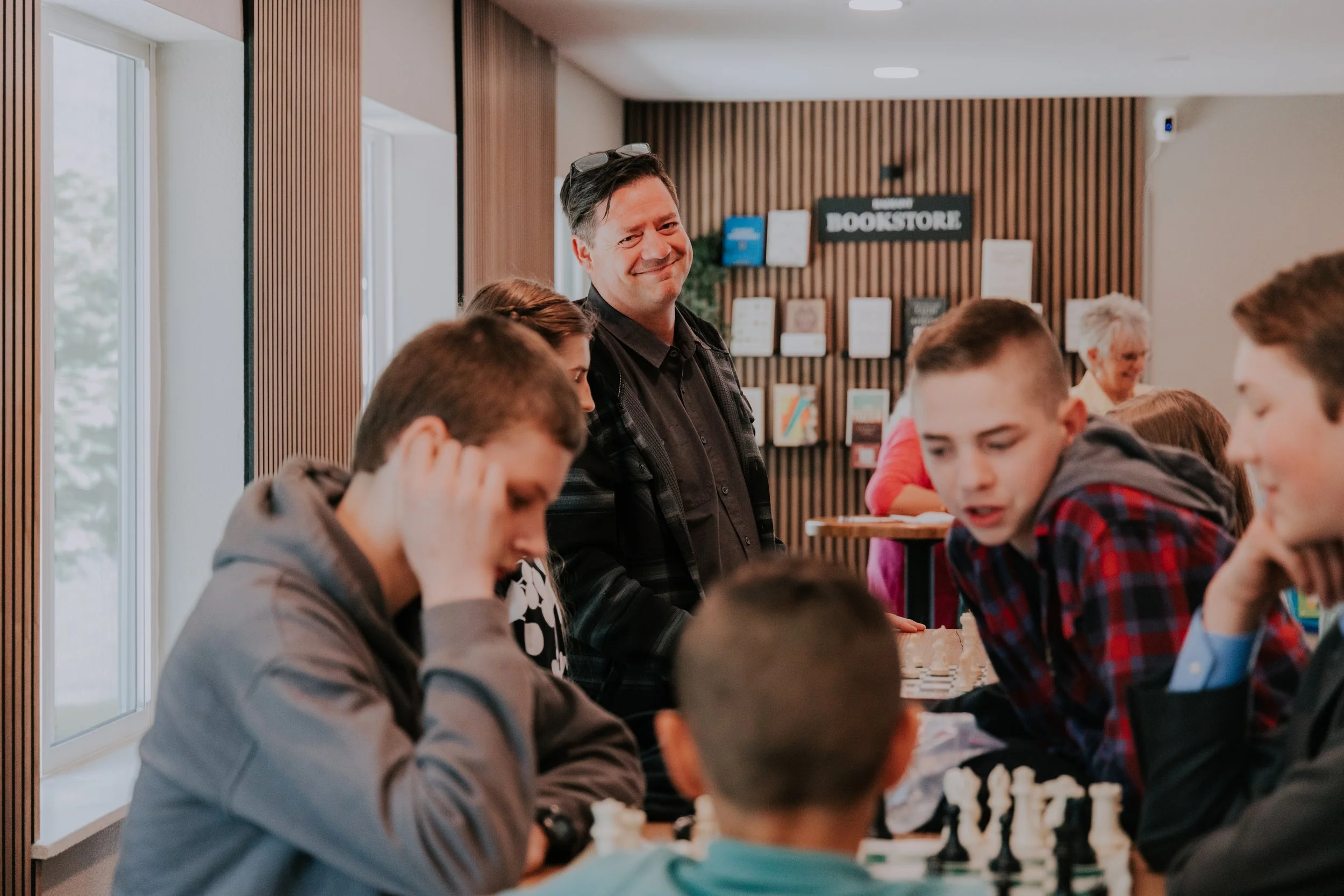 A man with glasses on his head smiling and standing among children playing chess in a library with a sign that reads 'BOOKSTORE' in the background.