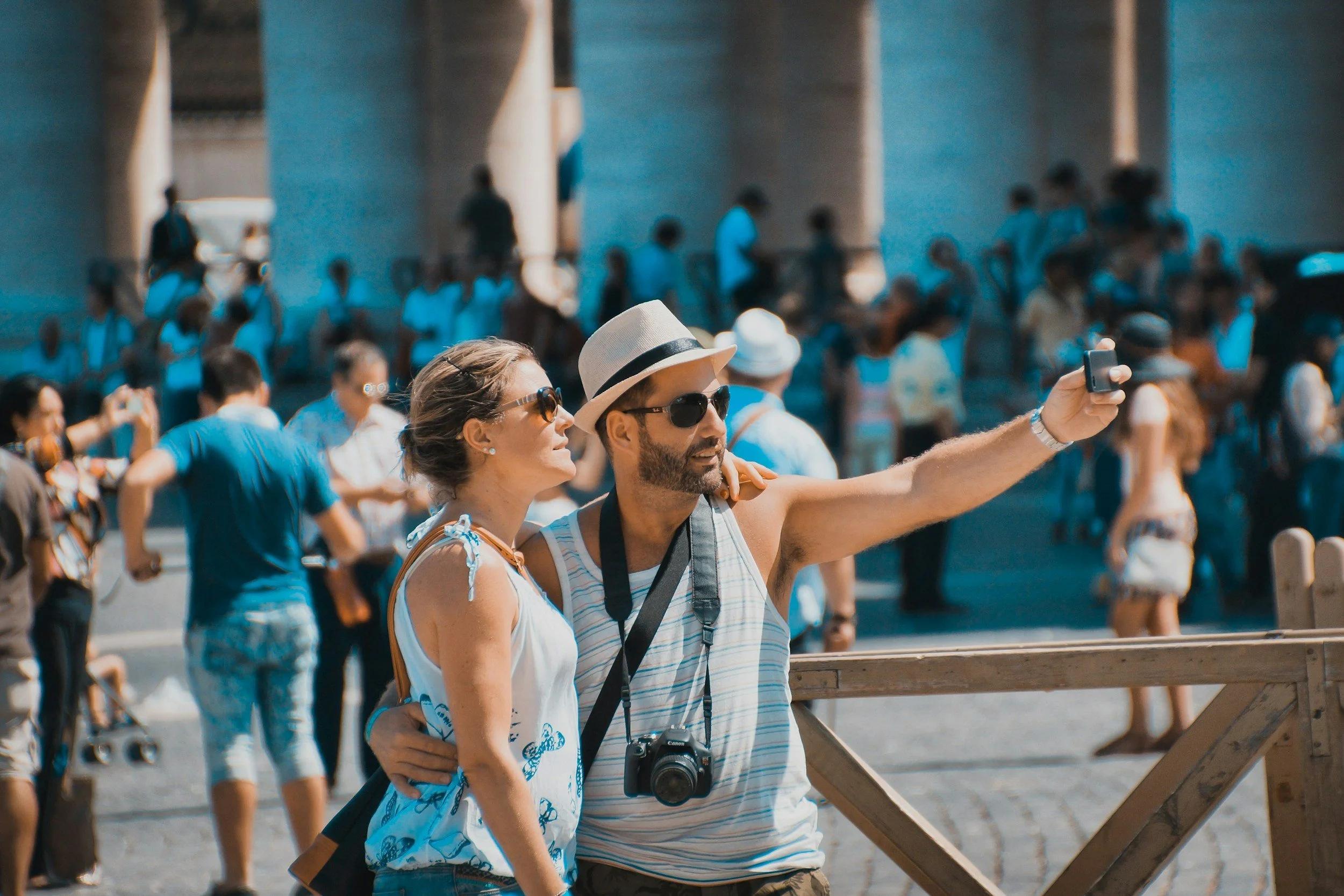 A man and woman taking a selfie at an outdoor event with many people in the background. The man is wearing a hat and sunglasses, the woman has sunglasses and curly hair. The crowd is gathered in a sunny area under a bridge or overpass.