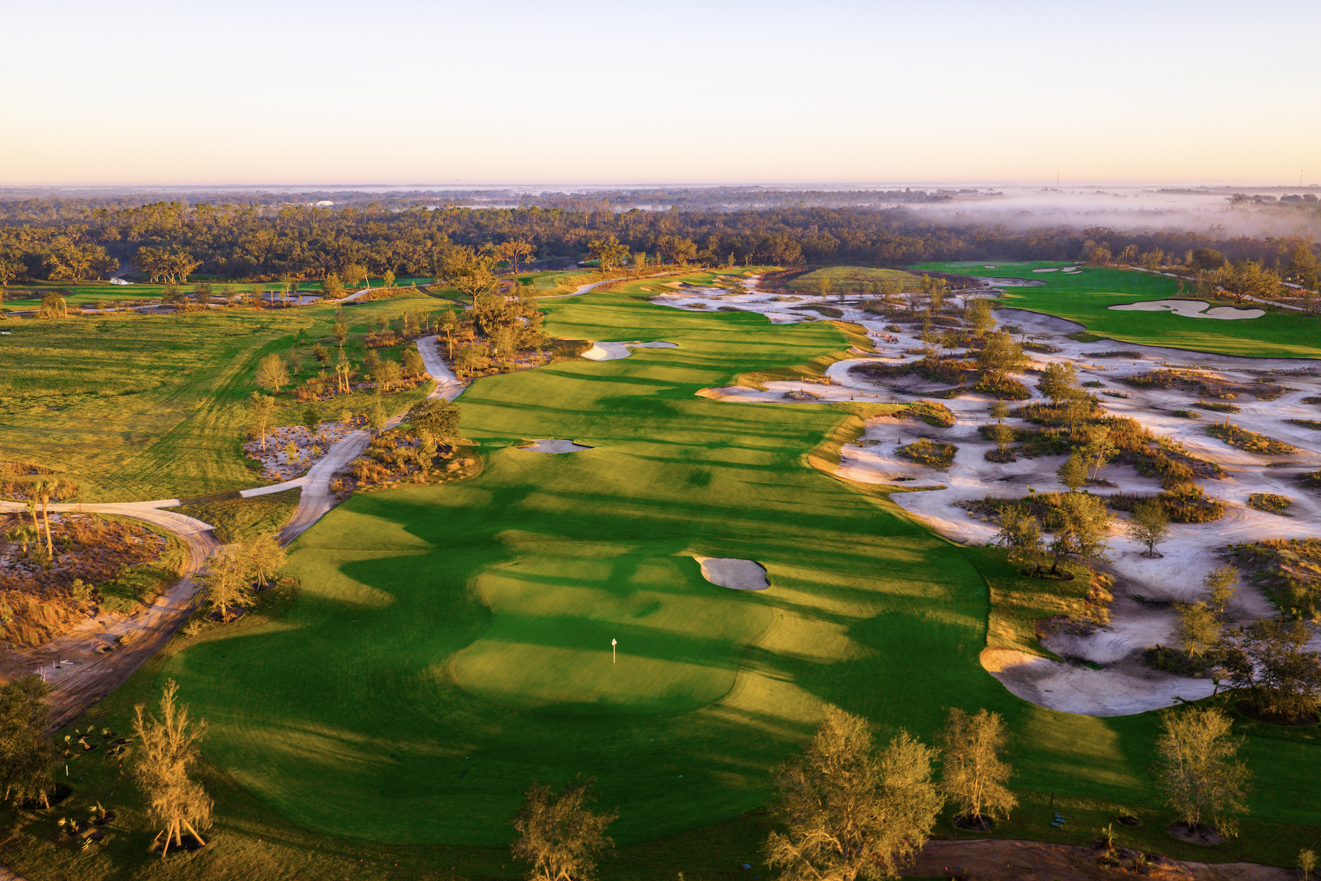 Aerial view of a golf course in the early morning, showing lush green fairways, sand bunkers, and surrounding trees under a clear sky with a horizon of distant trees.