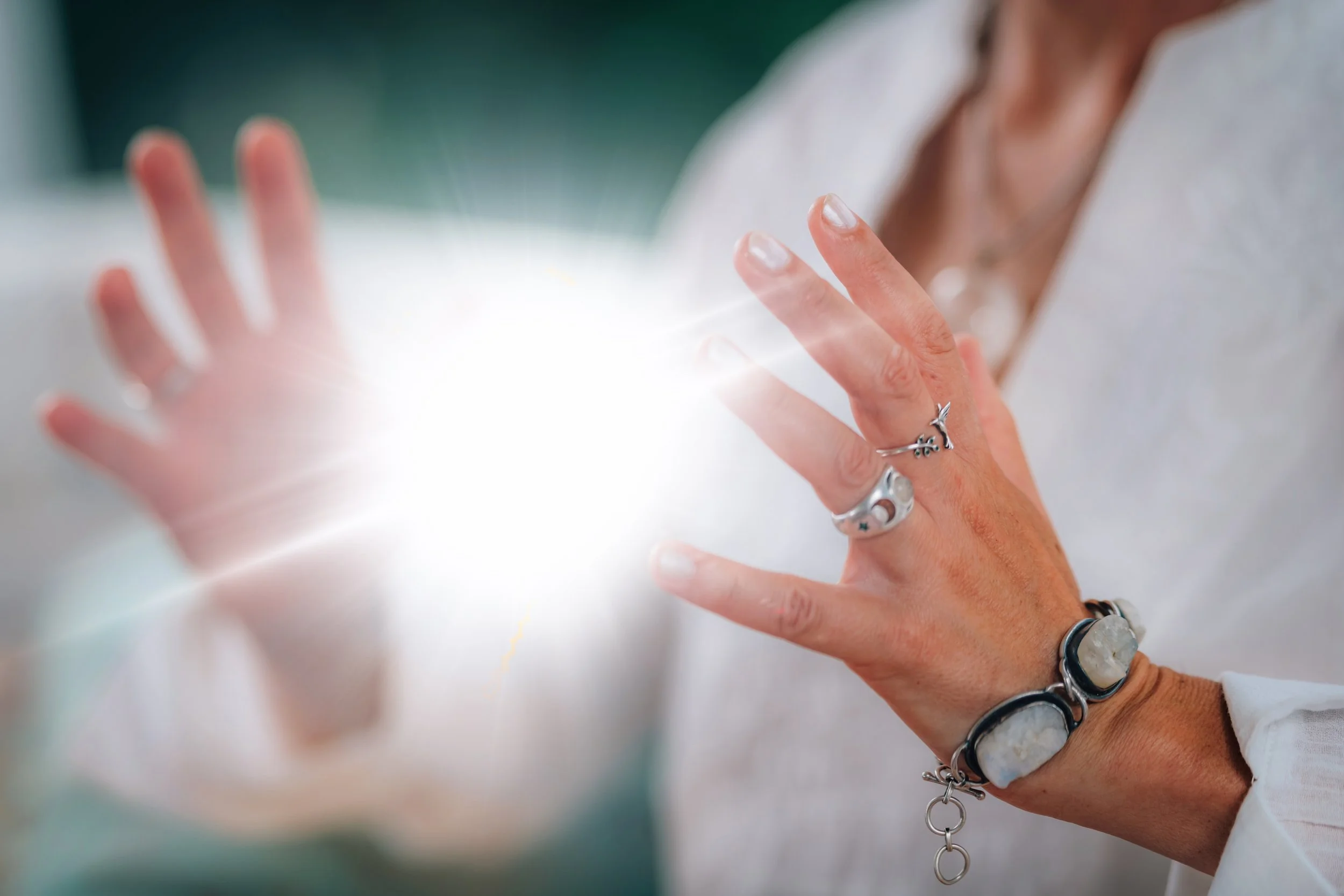 A person holding their hands near a bright, glowing light with objects on their fingers and wrist, wearing various rings and bracelets.