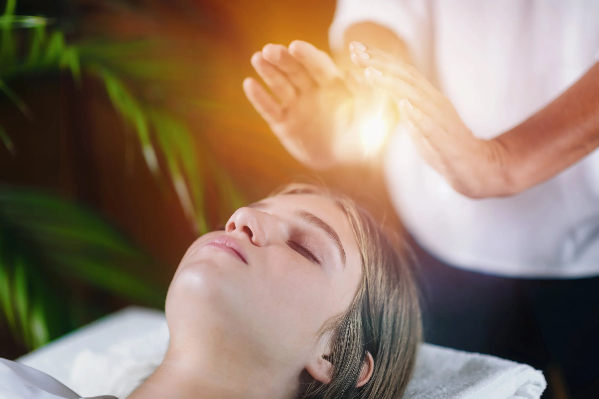 Person lying down with eyes closed, receiving a healing or energy treatment from hands above their face, with sunlight and green plants in the background.