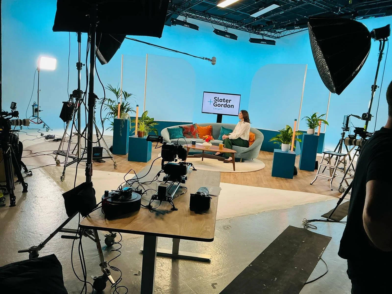 A television studio or set with a woman sitting on a sofa, surrounded by potted plants, with a monitor displaying 'Slater + Gordon Lawyers' behind her. The set includes professional lighting and camera equipment.