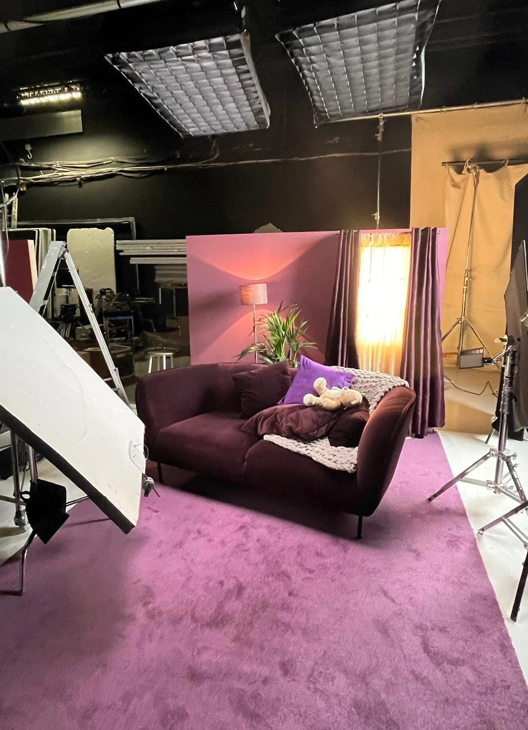 Photo of a TV studio set with a purple sofa, pillows, a teddy bear, a blanket, a plant, a lamp, a window with curtains, and professional lighting equipment.