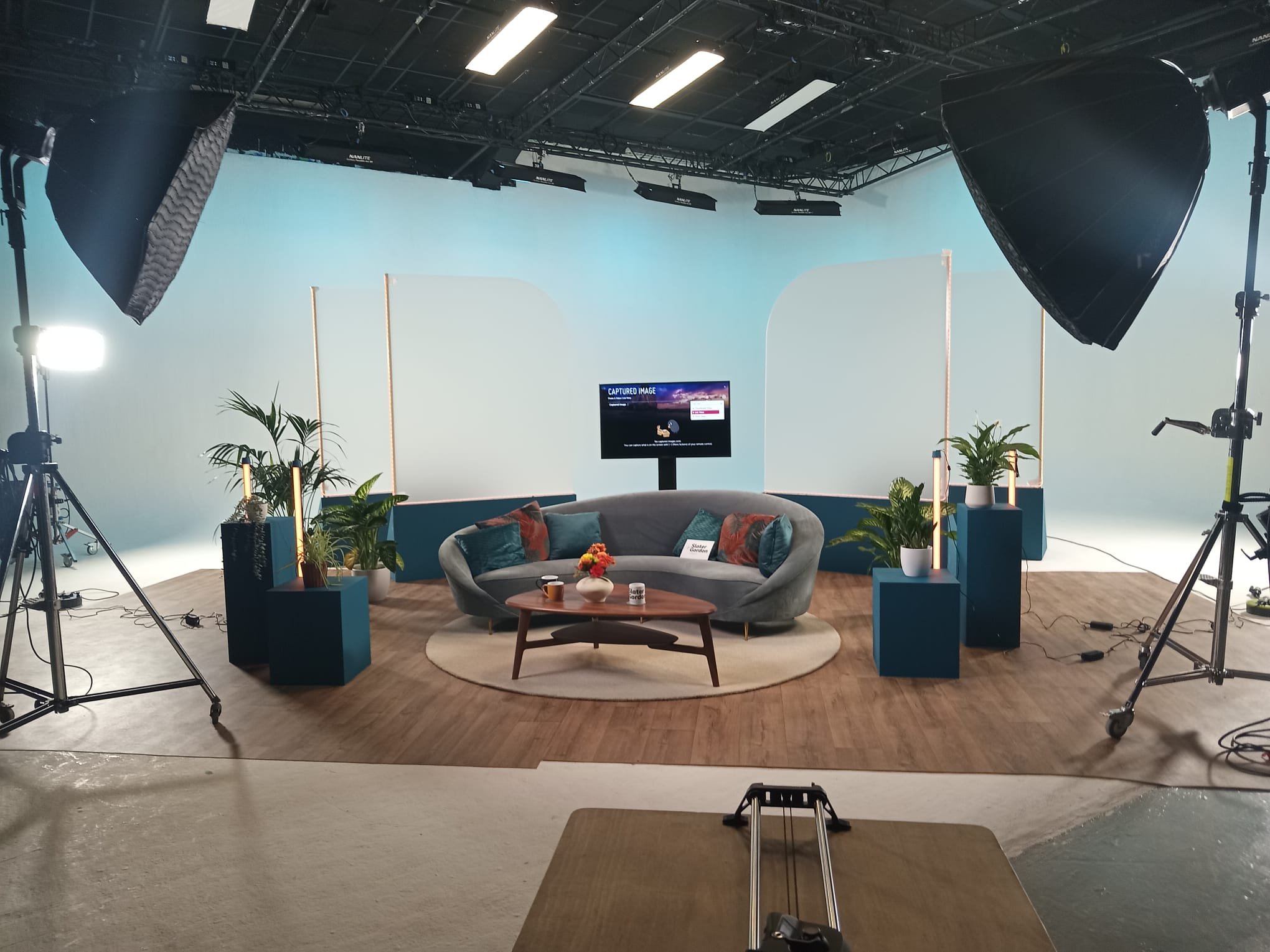 A television studio set with a gray curved sofa, a small wooden coffee table with a flower vase, several cushions, and potted plants. The background is a light blue screen, with lighting and camera equipment visible.