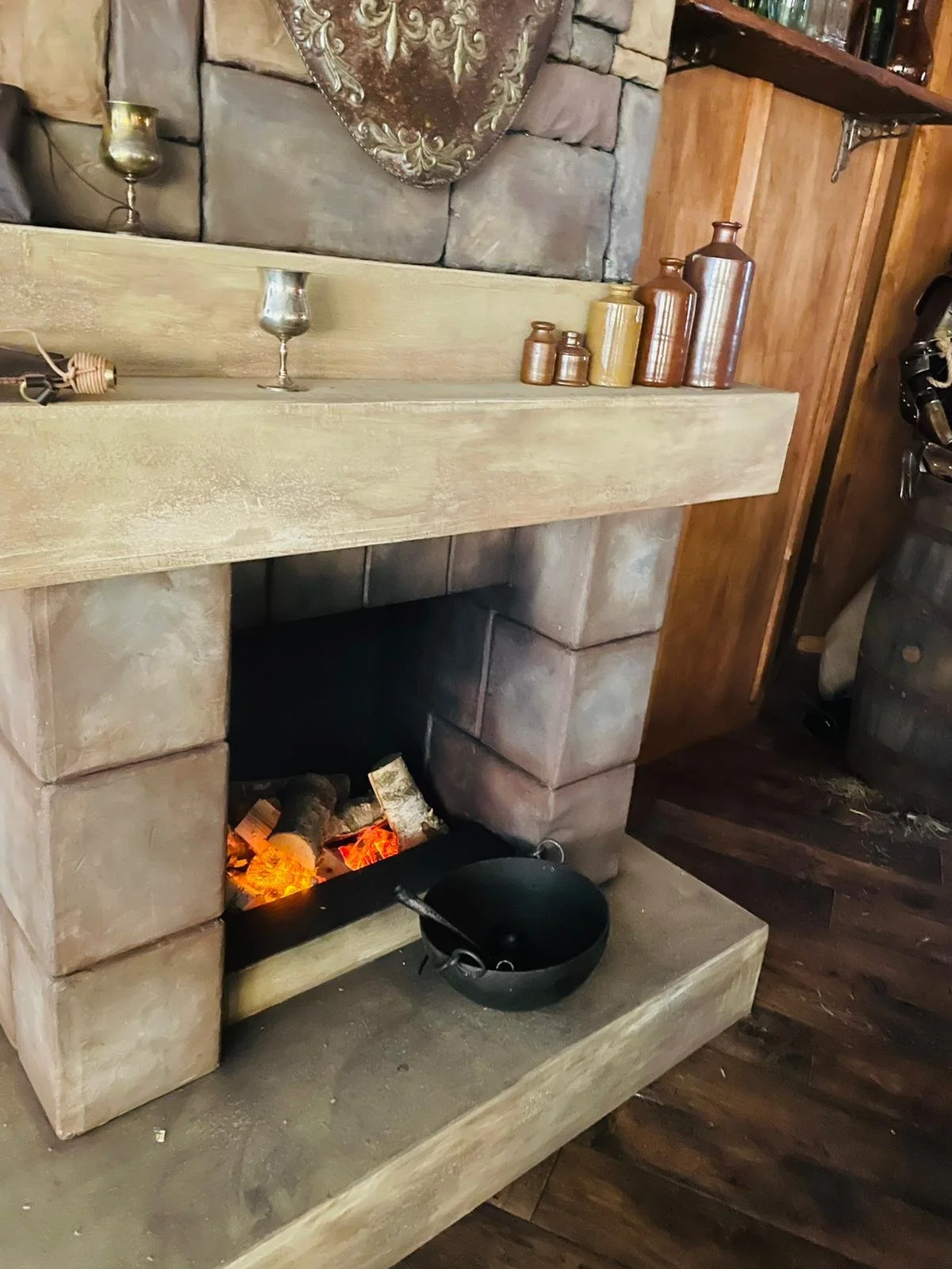A cozy fireplace with a stone surround and uncooked logs inside. A black pot hangs on the hearth and a wooden mantel above holds decorative bottles and candle holders. The room has wood paneling and a stone wall.