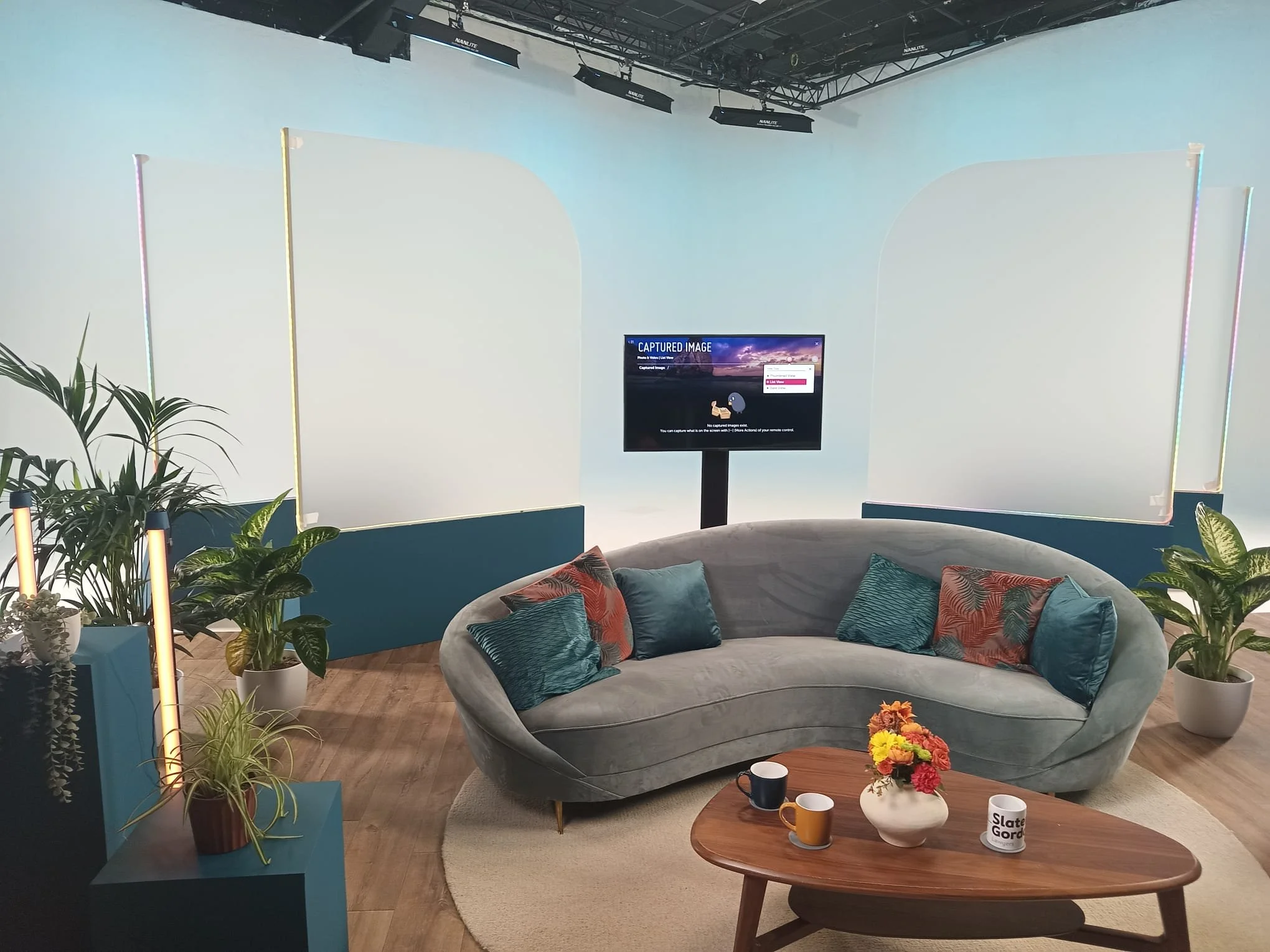 Modern TV studio with a curved grey velvet sofa, colorful cushions, a wooden coffee table with flowers and mugs, green potted plants, and a large flat screen monitor on a stand. The background features white screens and a light blue backdrop.