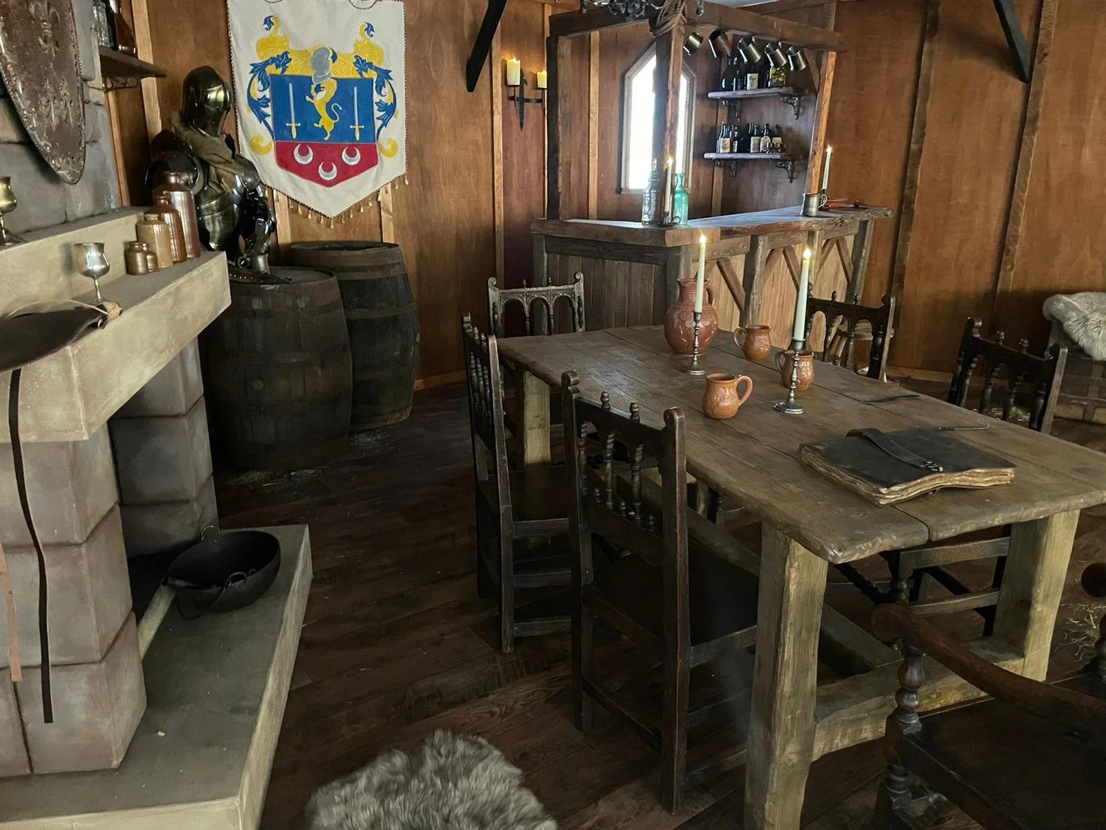 A rustic interior scene with a wooden dining table set with three candles, a large jar, and smaller clay vessels. The room features wood-paneled walls, a shield or banner with a heraldic design hanging on the wall, two wooden barrels, and a small bar area with shelves holding bottles and glasses. A shield or statue of armor is placed on the left side near the barrels. The lighting is warm, with candles and a wall-mounted candle holder.