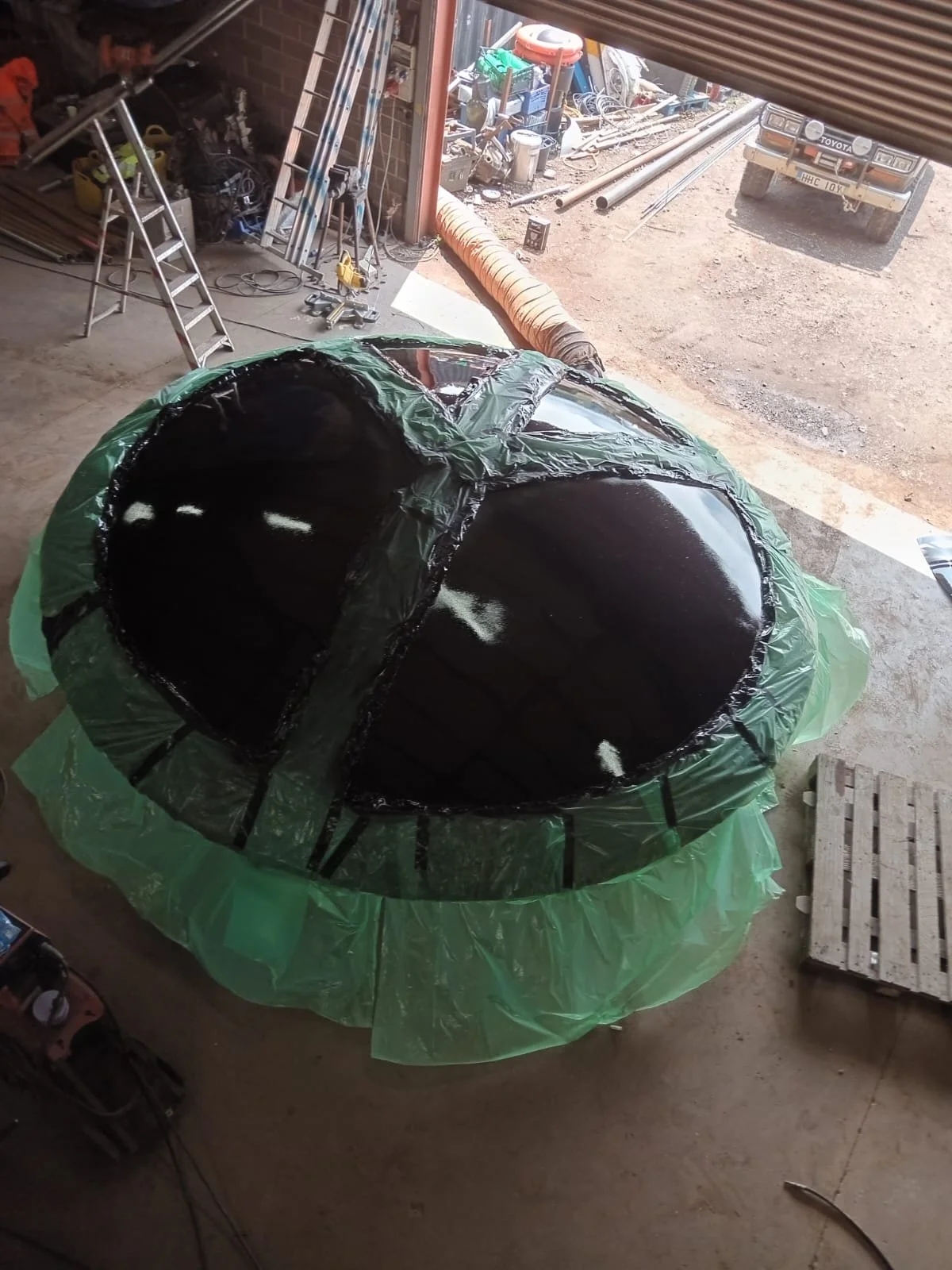 A workshop with a large spherical object covered in black material and surrounded by green plastic sheeting. The object appears to be under construction or repair, with tools, ladders, and equipment scattered around.