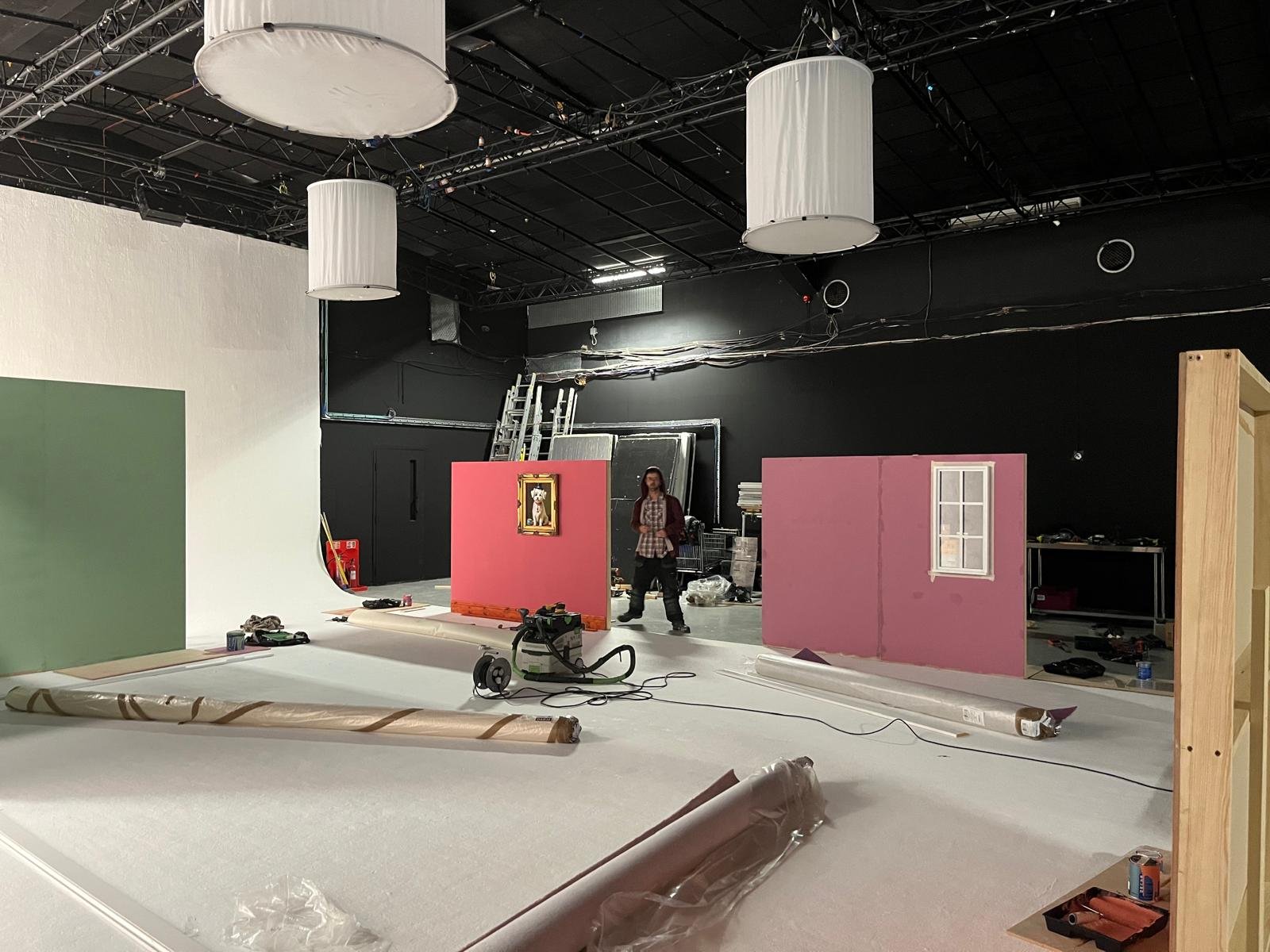 Set construction with partly painted walls, one green and one pink with a window, in a studio with hanging lights and equipment, a person standing near the pink wall.