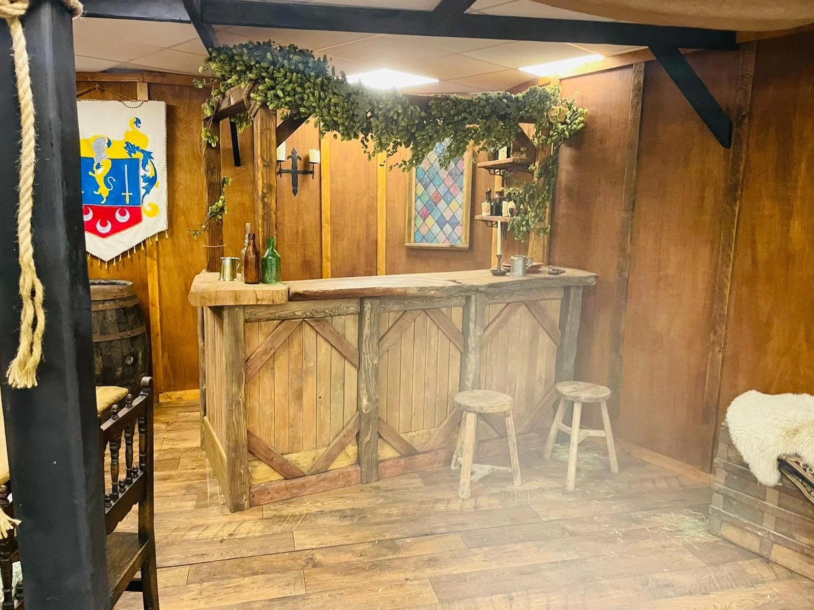 A rustic wooden bar with two stools in a warmly lit, medieval-style room decorated with shields, banners, and greenery.