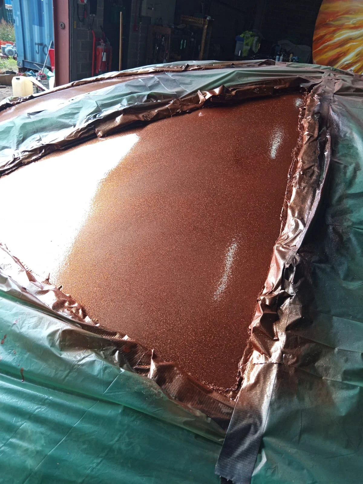 A large container filled with a shimmering, metallic rose gold or copper-colored material, possibly resin or paint, lined with black plastic or foil.