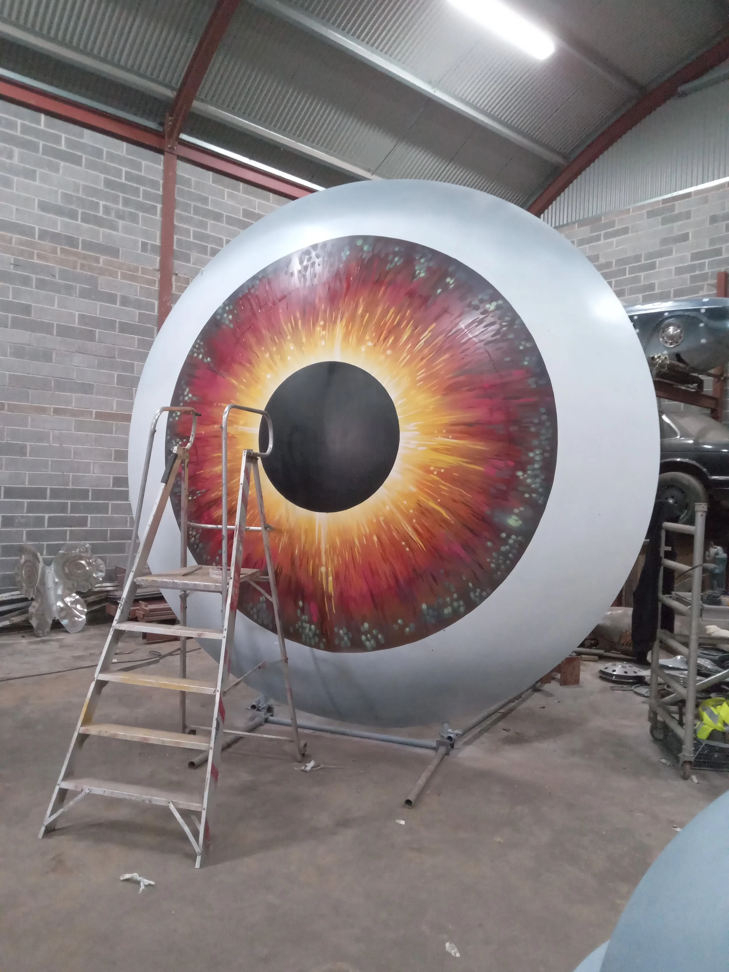 Large scale art installation of a giant human eye with a colorful iris pattern, located in an industrial workshop with brick walls and metal roof.