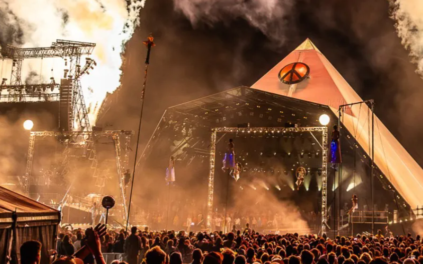 A large outdoor concert stage with performers and aerial performers, pyrotechnics and smoke effects, and a crowd of spectators at night.