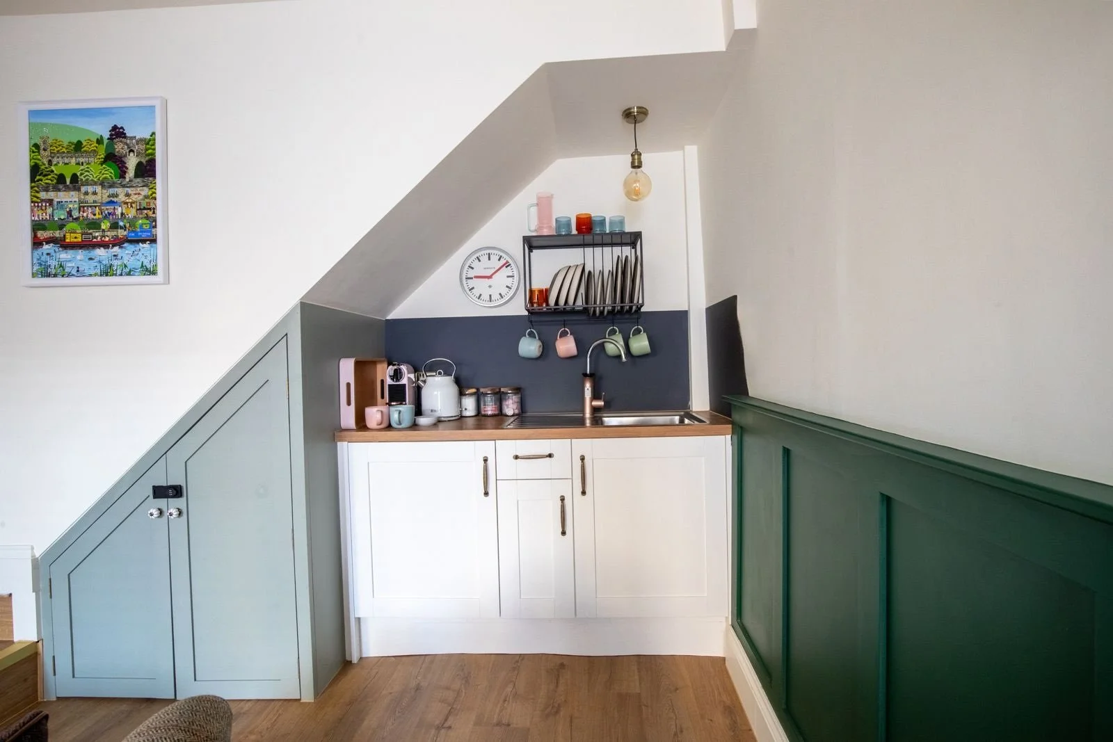 Small kitchen under a sloped ceiling with white and green walls, a wooden countertop, and hanging cups above a sink.