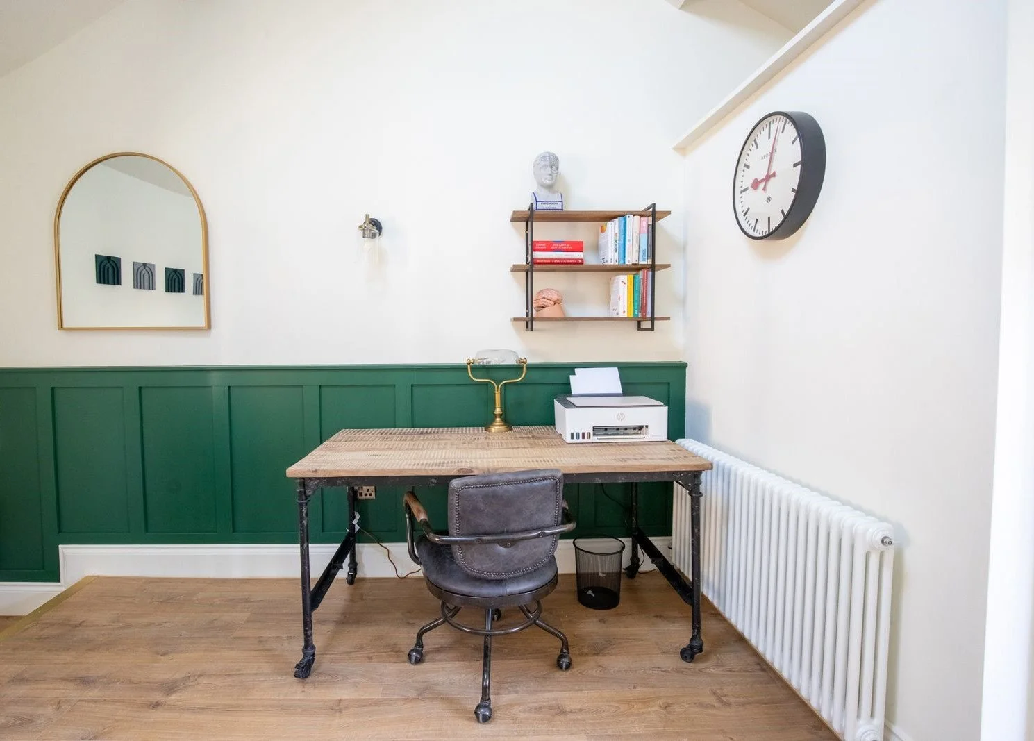 A small home office with a wooden desk, vintage black swivel chair, white printer, radiator, and wall decor including a large wall clock, mirror, and shelves with books and ornaments.